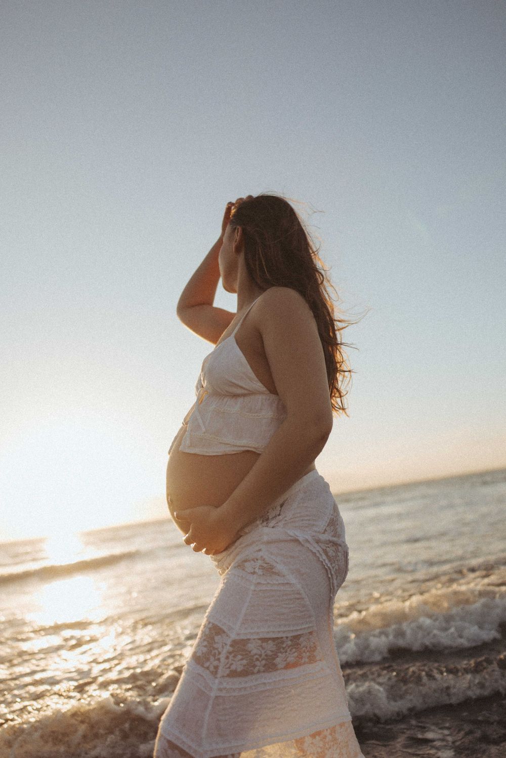 Pregnant woman in white lace dress cradling her baby bump on a sunlit beach at sunset in Raglan New Zealand.
