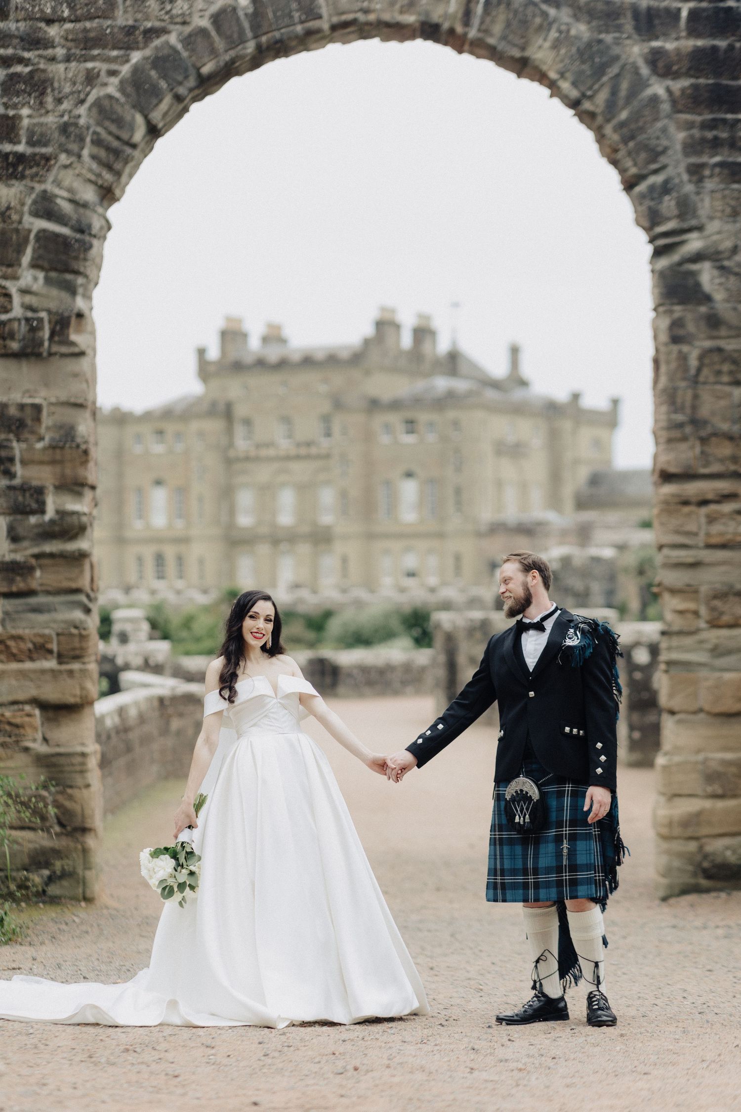 Bride and groom in tartan kilt holding hands under a stone arch with a castle backdrop.