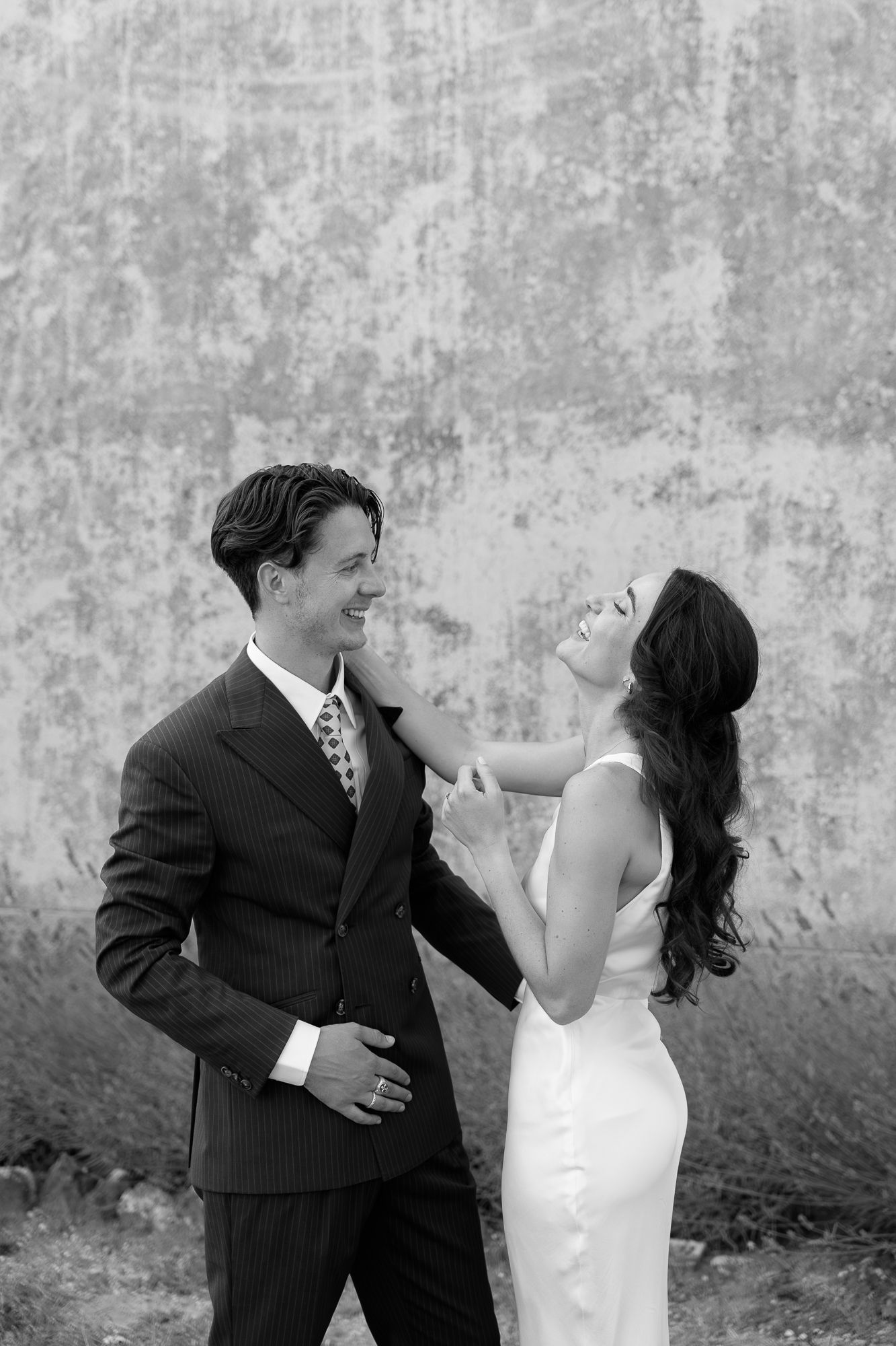 Black and white photo of a smiling bride and groom in elegant attire sharing a joyful moment against a rustic wall.