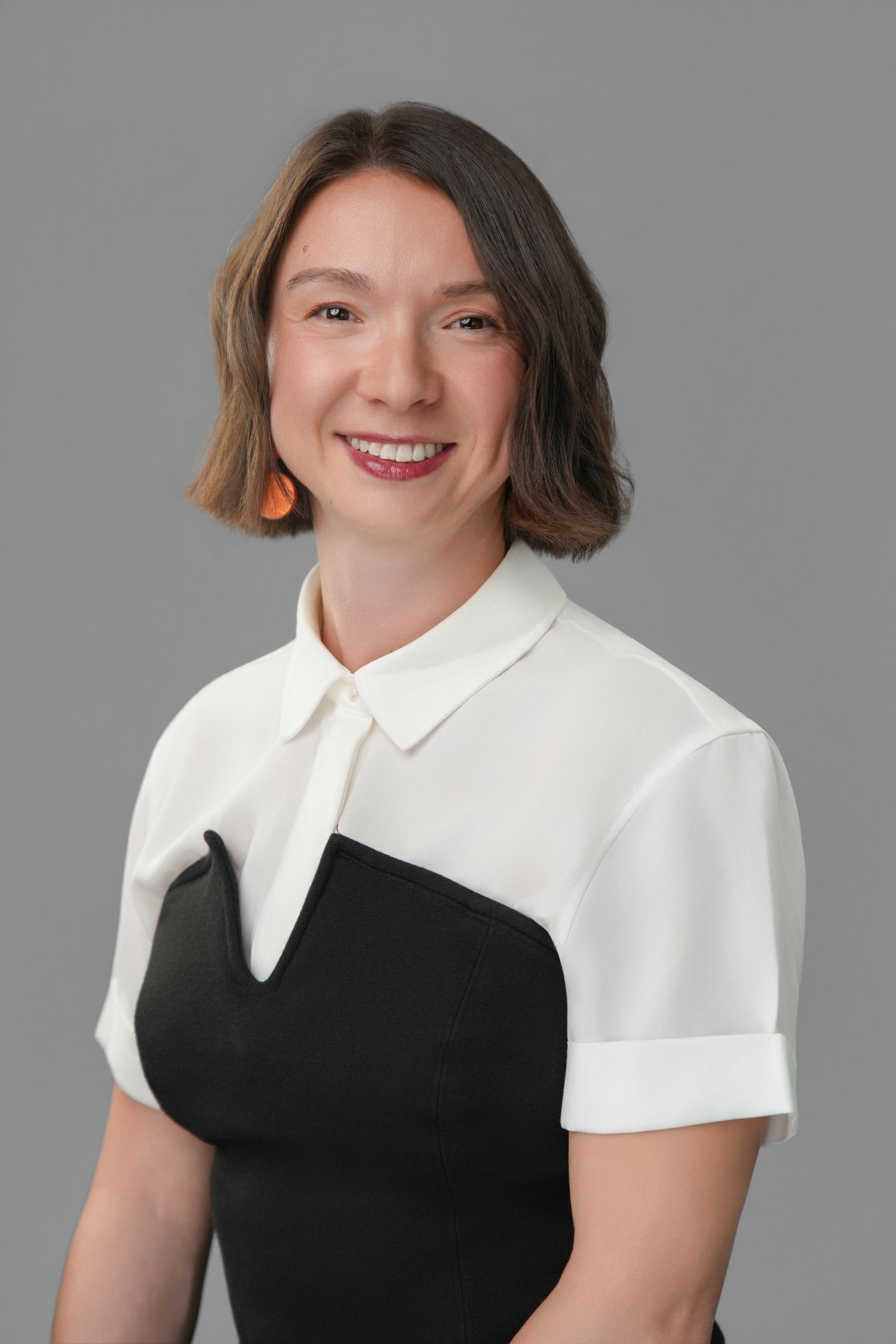 Professional woman smiling in black and white top with orange earrings against gray background.