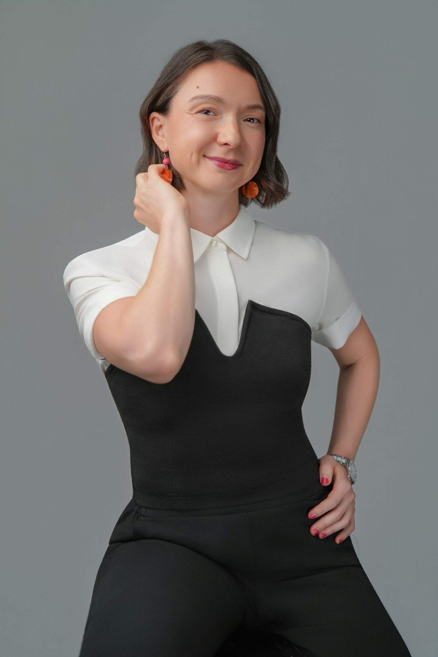 A smiling woman in a white blouse and black vest poses confidently against a gray background, wearing orange earrings.