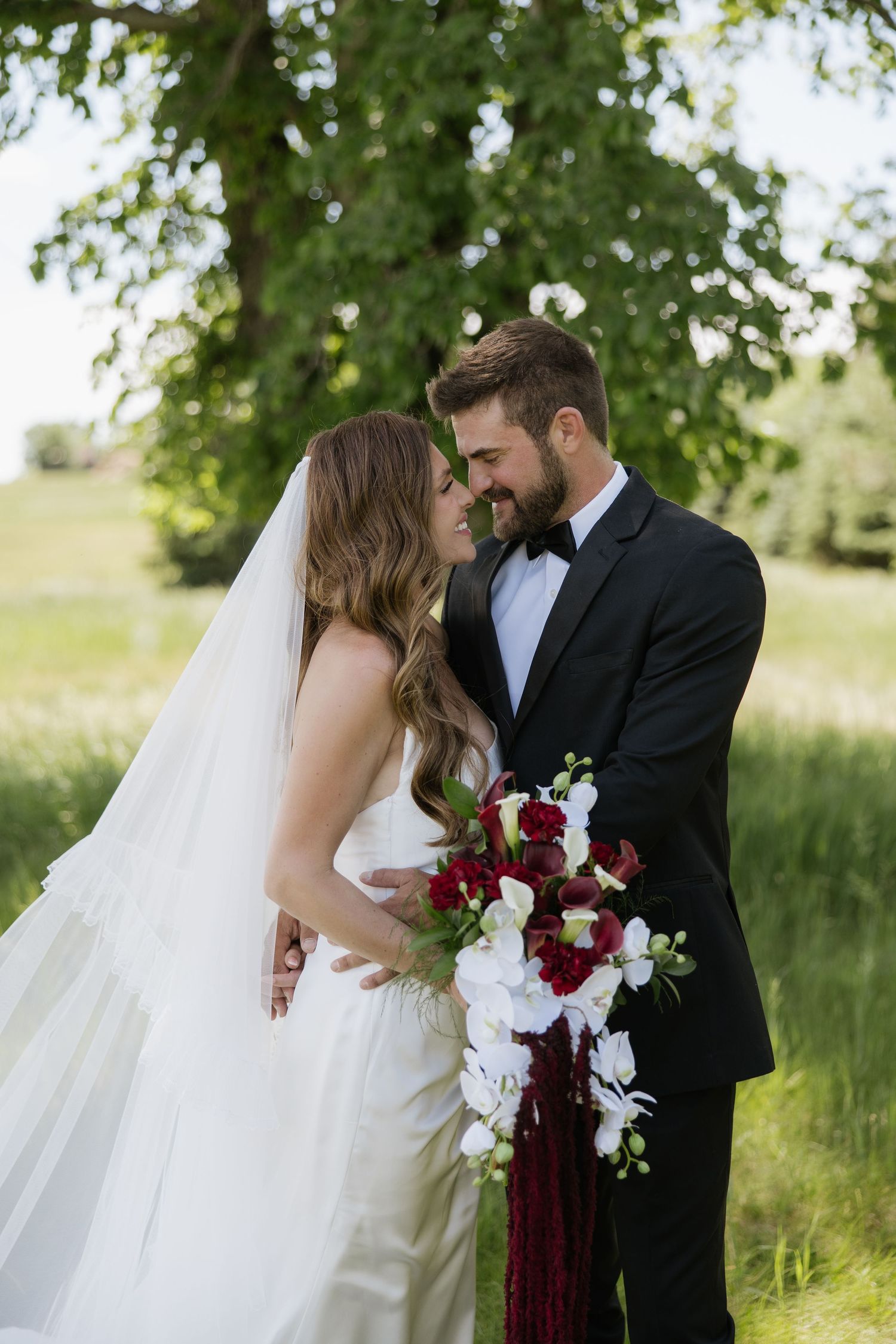 Bride and groom share a romantic moment outdoors, she holds a red and white cascading bouquet, both in elegant wedding attire.