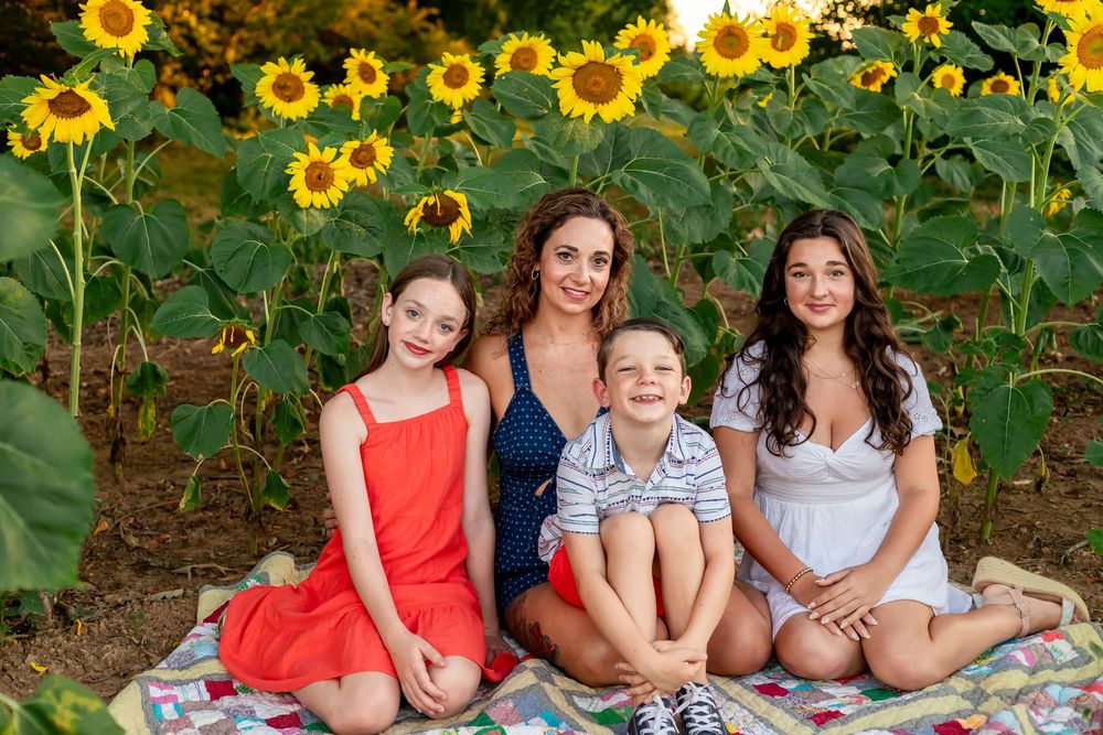 Family of four smiling in a sunflower field, sitting on a quilt outdoors on a sunny day.