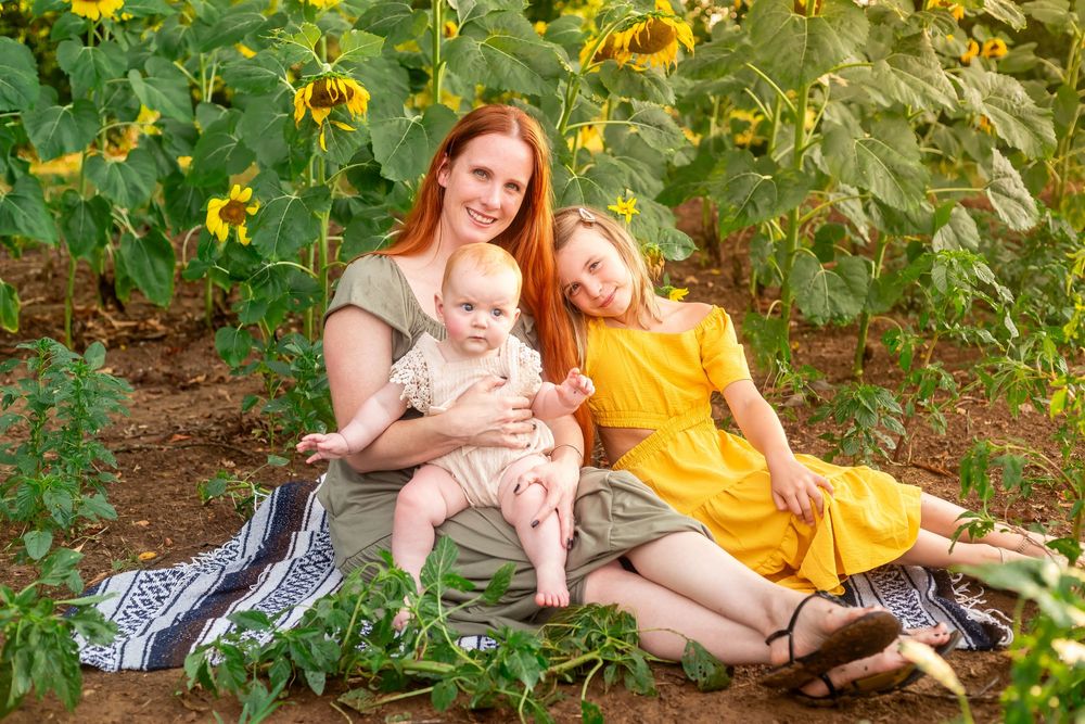A red-haired mother sits with her baby and young girl in yellow dress among sunflowers in a sunny field.