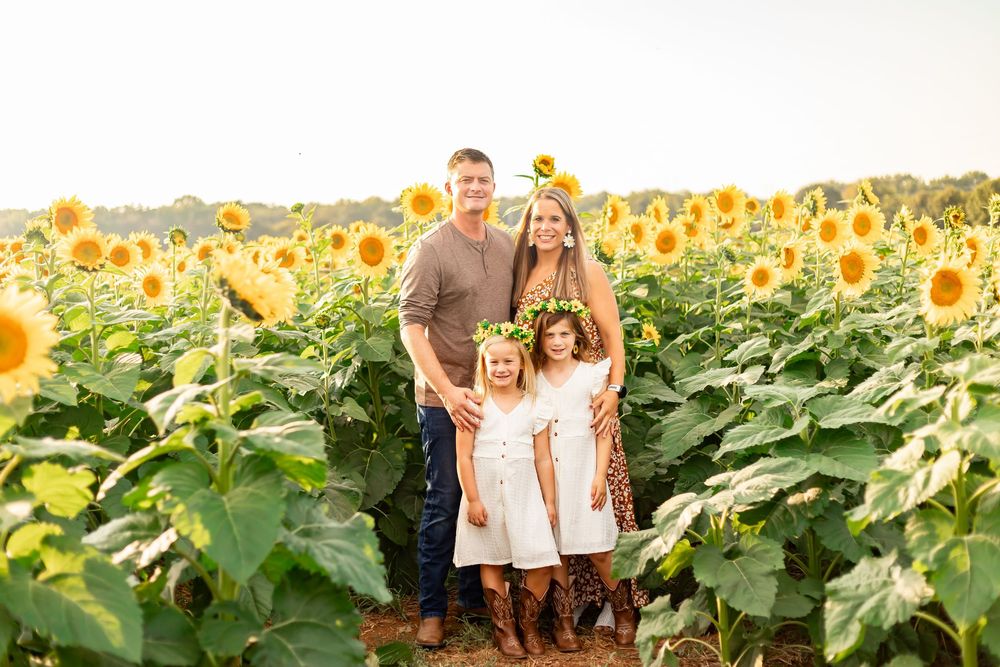 Family of four posing in a sunflower field, two girls wearing white dresses and flower crowns, parents smiling outdoors.