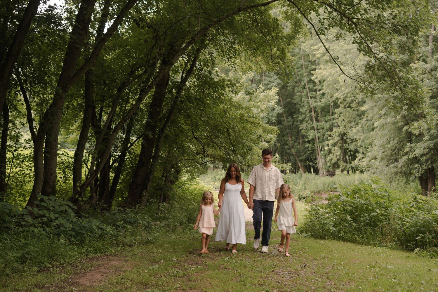 Family of four walking hand-in-hand on a lush green forest path surrounded by tall trees.