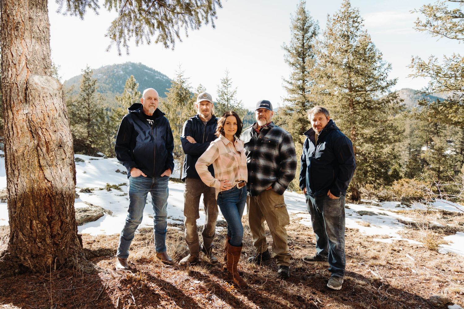 Five people posing outdoors in a snowy mountain forest with evergreen trees and snow-dusted hills in the background.