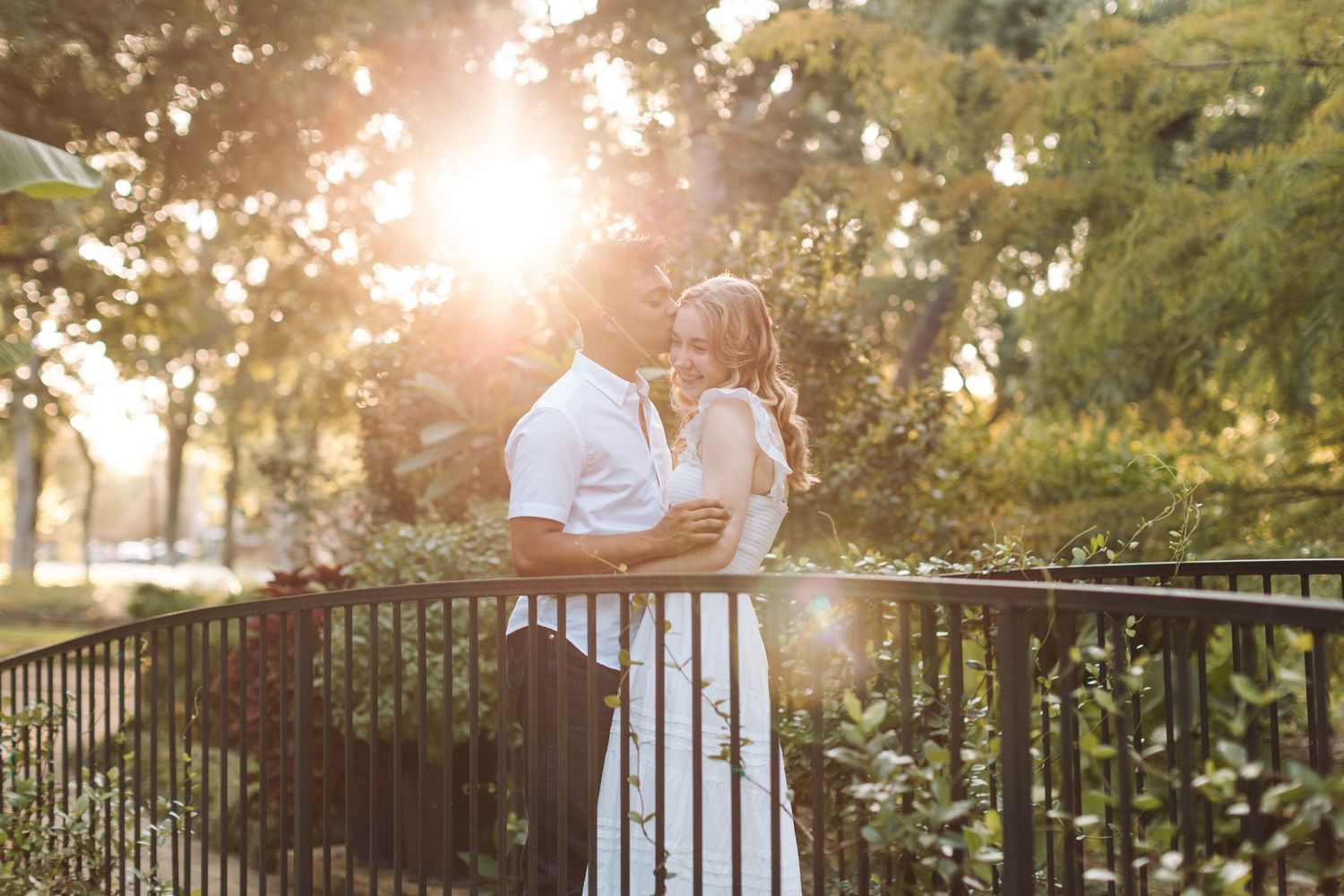 Couple embracing on a bridge at golden hour, surrounded by lush green trees with warm sunlight glowing behind them.