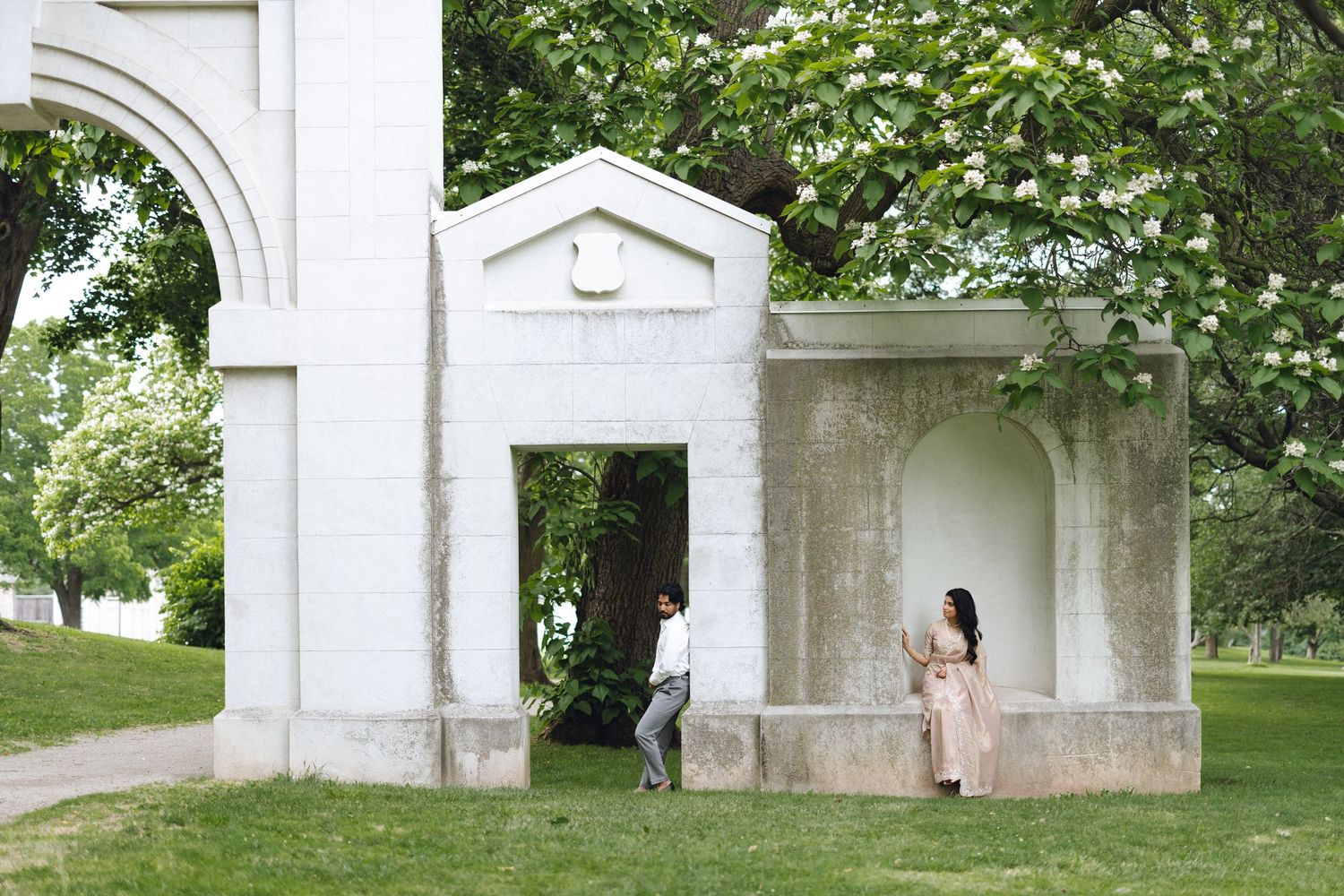 Couple posing near a white stone archway structure in a lush green park with blooming trees.