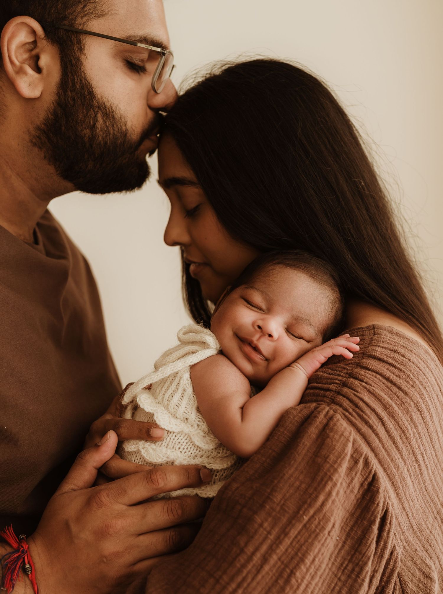 Parents tenderly holding their smiling newborn baby wrapped in a white knit blanket in a warm, intimate portrait.