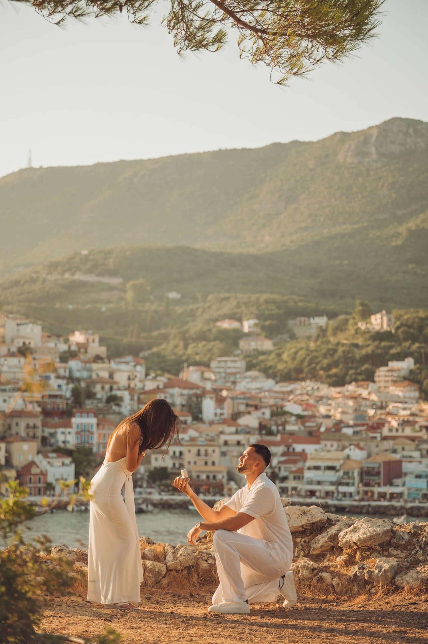 Man proposing to woman in white dress on hilltop overlooking a scenic Mediterranean coastal town at sunset.
