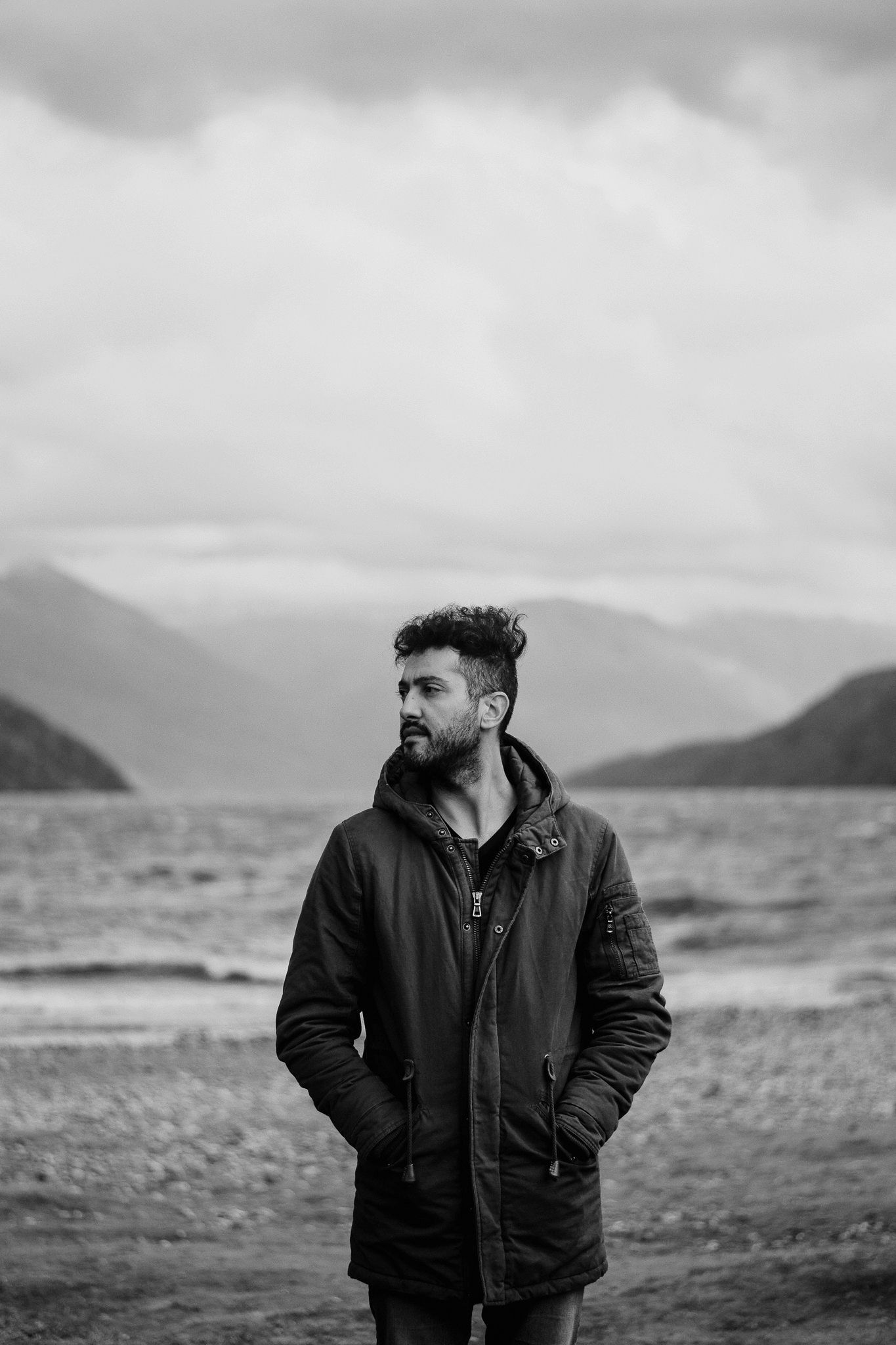 Black and white portrait of bearded man in winter jacket standing by a lake with mountains in background.