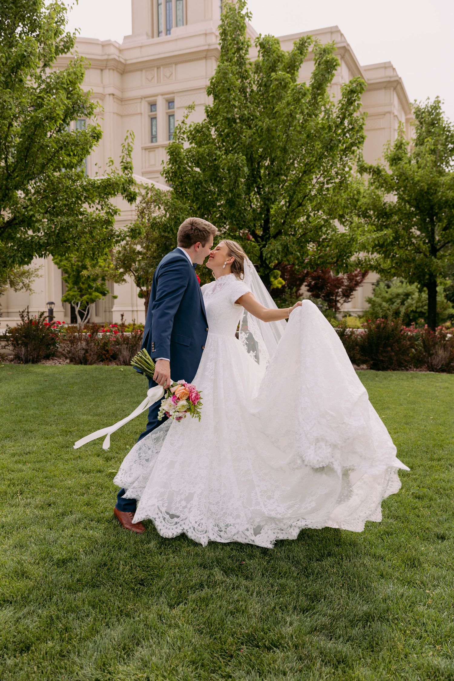 Bride and groom hold hands on steps before a grand LDS temple with a fountain and colorful flowers.