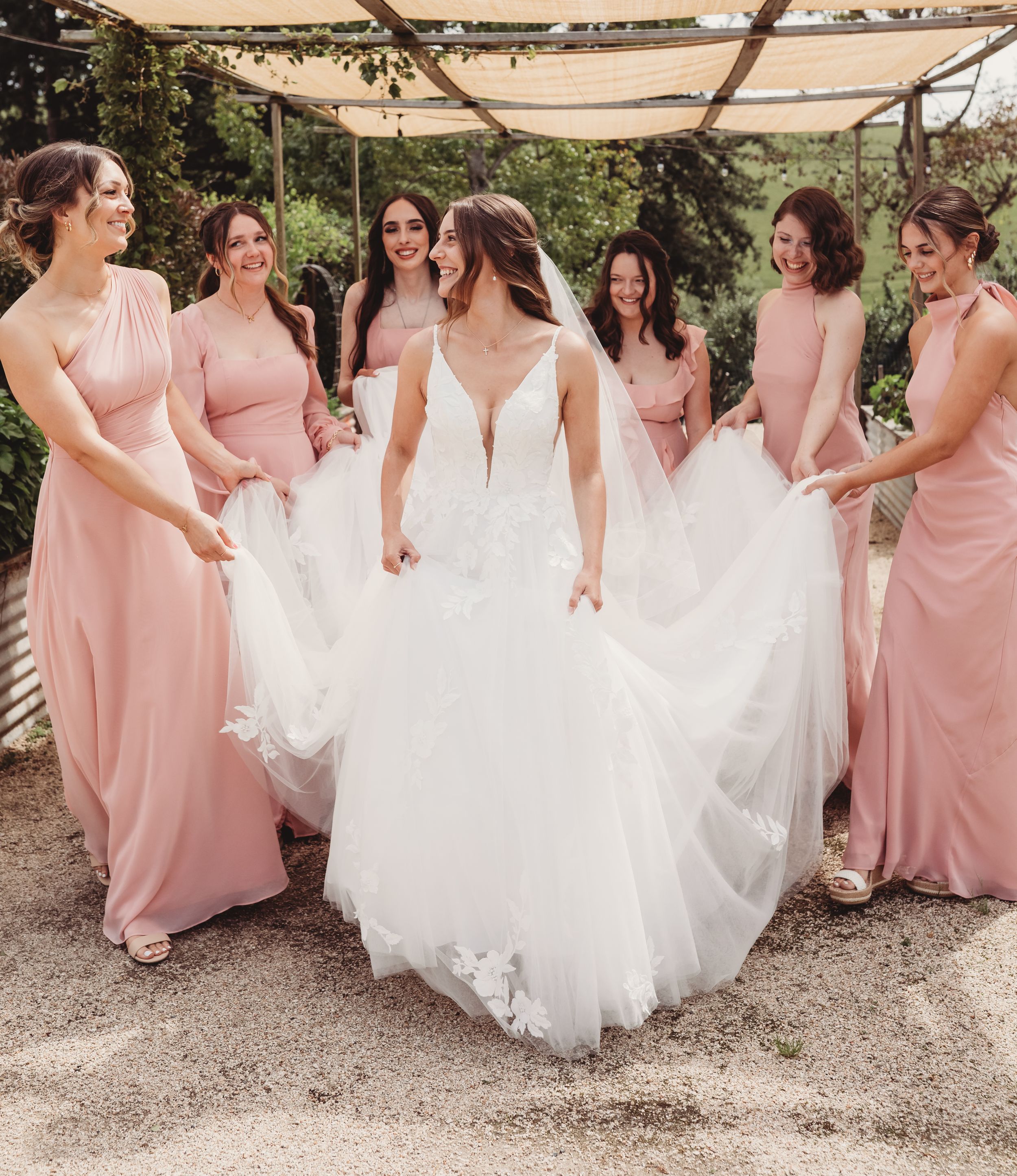 Bride in white gown laughs with six bridesmaids in pink dresses holding her train outdoors.