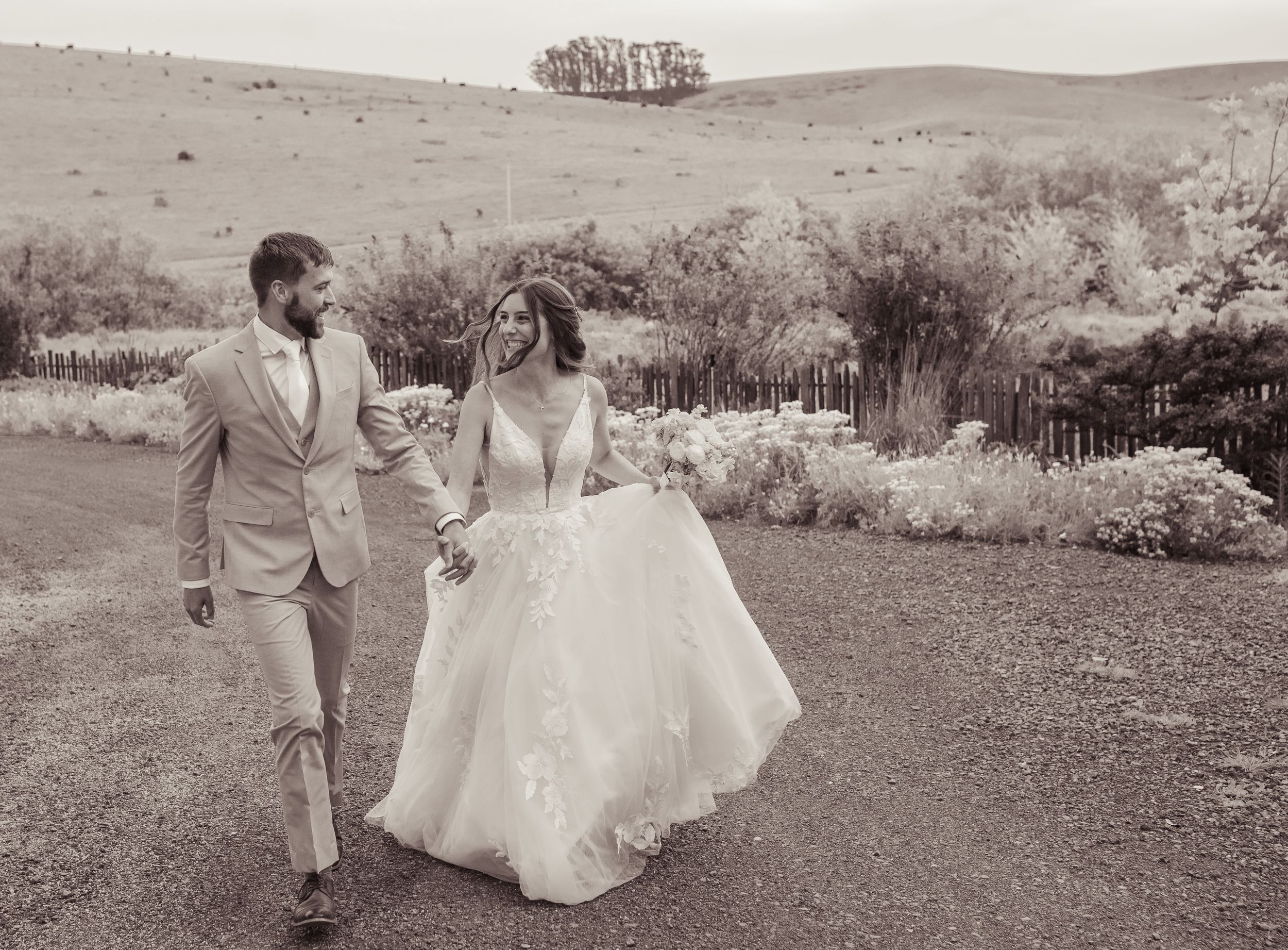 Black and white photo of bride and groom holding hands walking along garden path.
