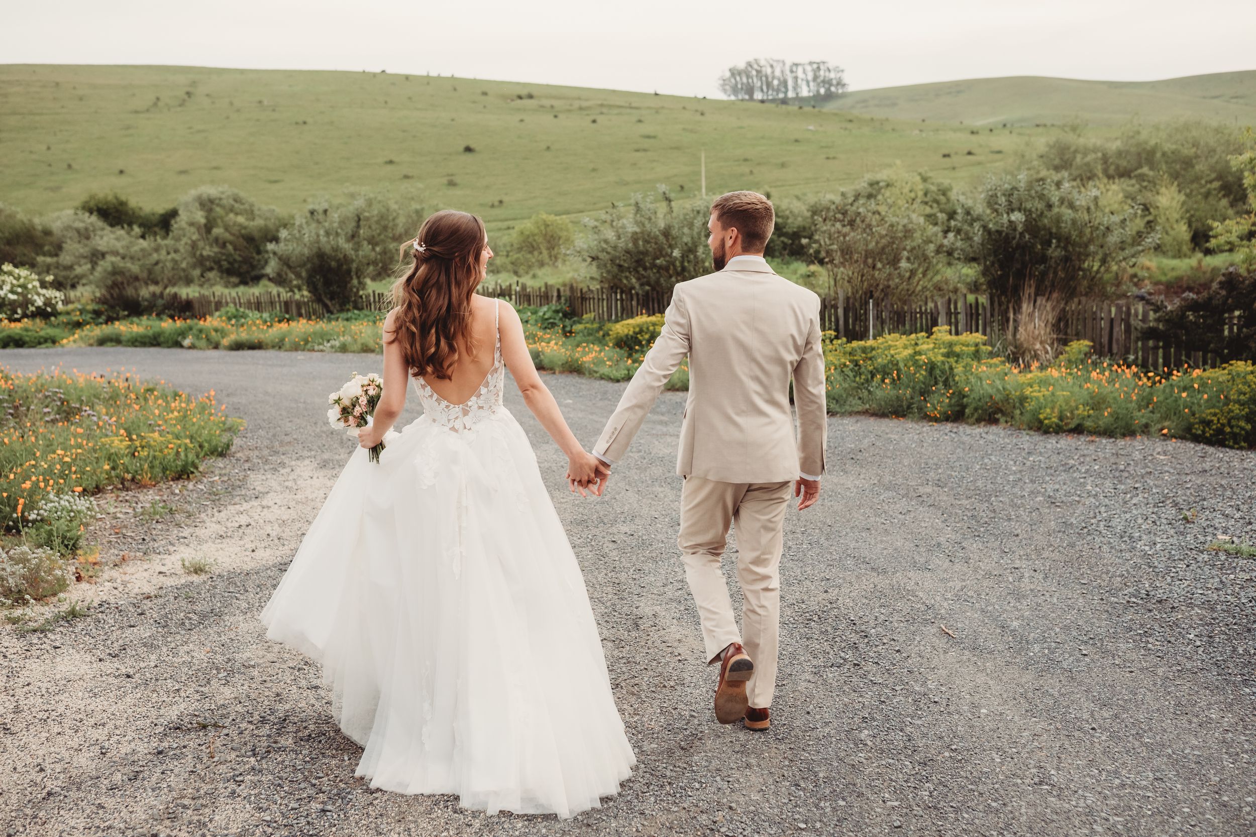 Bride and groom walking hand in hand on a gravel path surrounded by lush green hills on their wedding day.