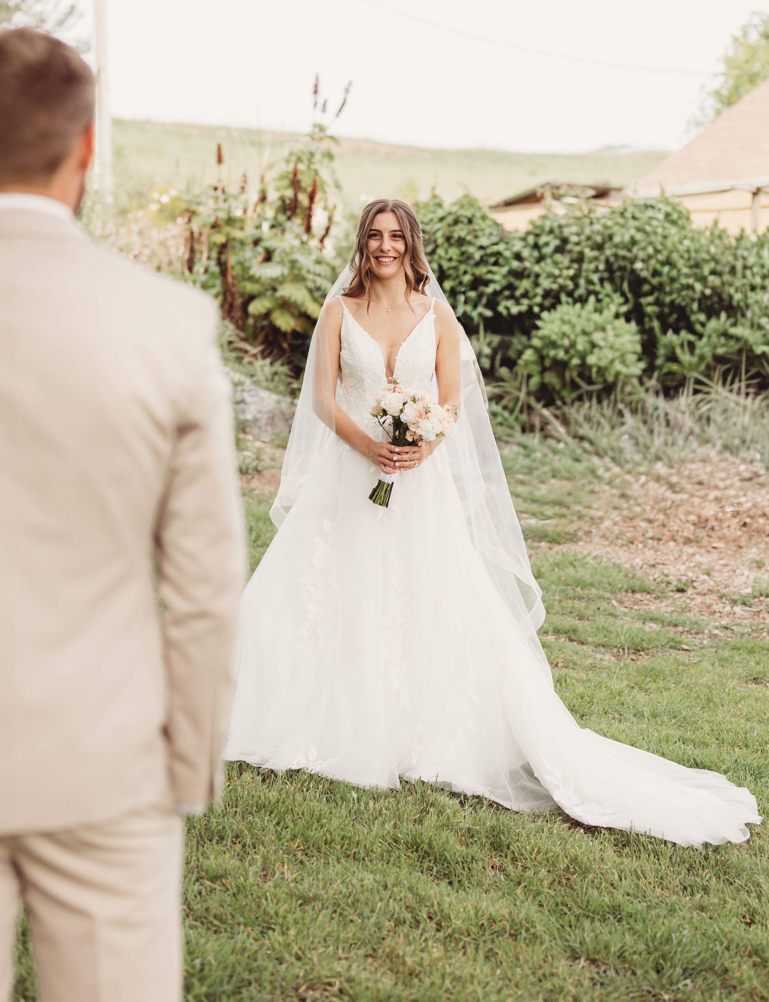 Bride in elegant white gown with veil holding bouquet smiling at groom during first look outdoor wedding.