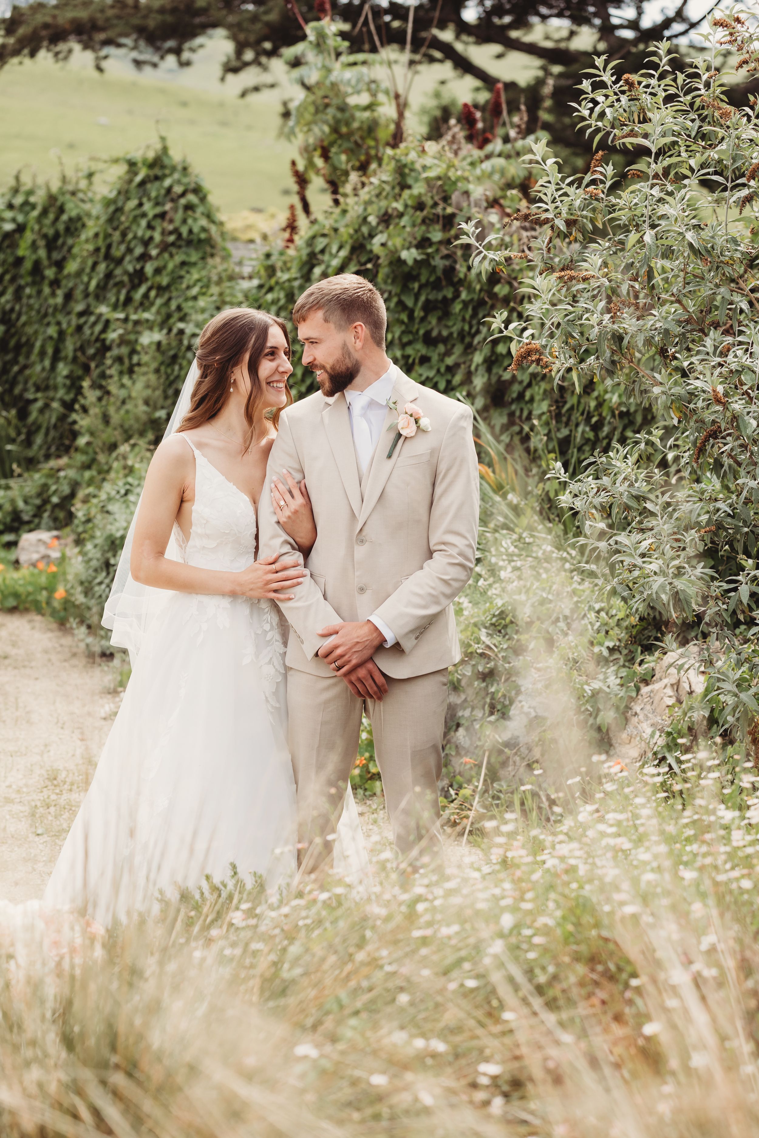 Bride and groom share a tender moment outdoors, surrounded by lush greenery, bride in white gown, groom in beige suit.