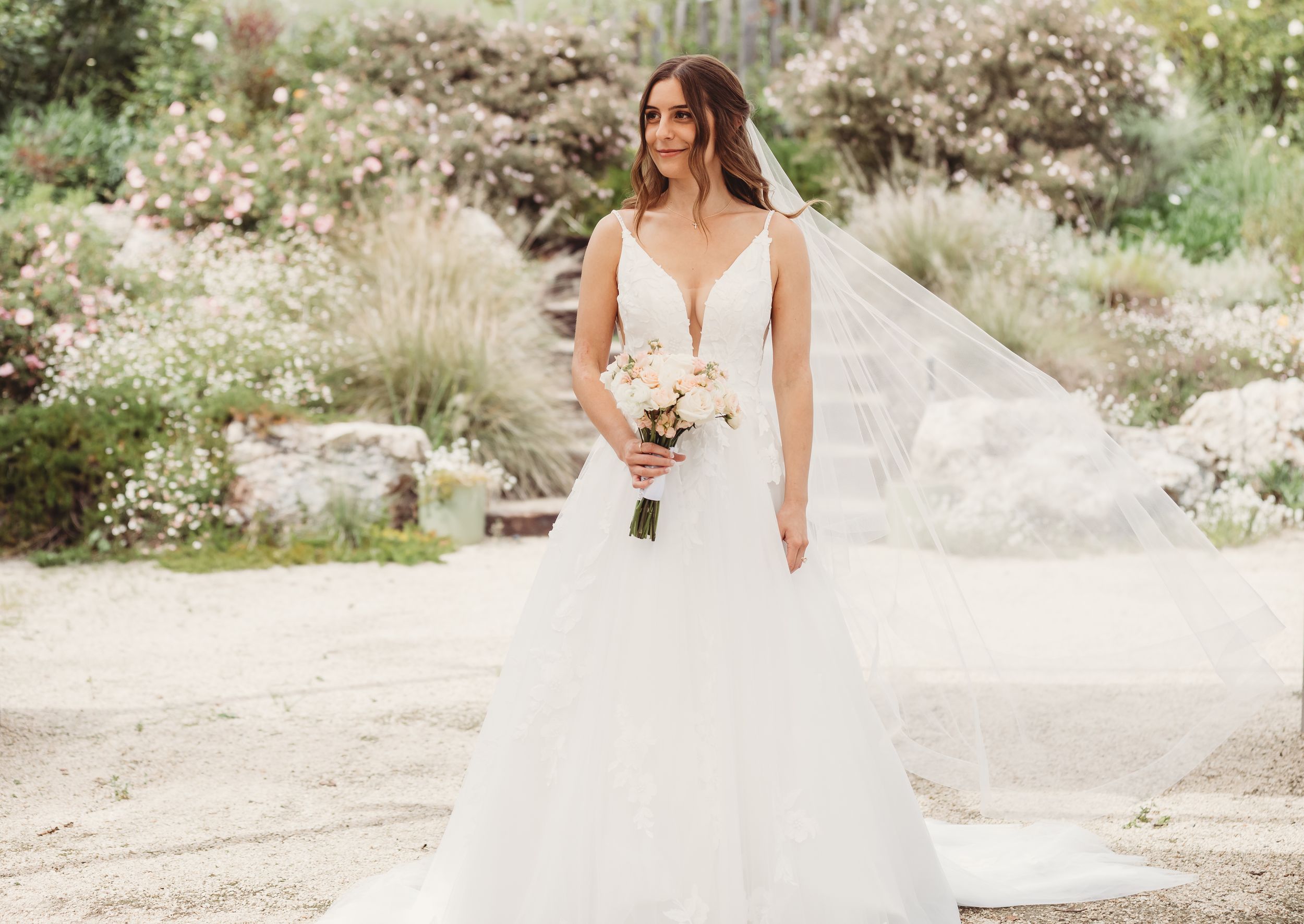 Bride in elegant white ball gown with deep V-neckline and flowing veil holds bouquet in a garden.