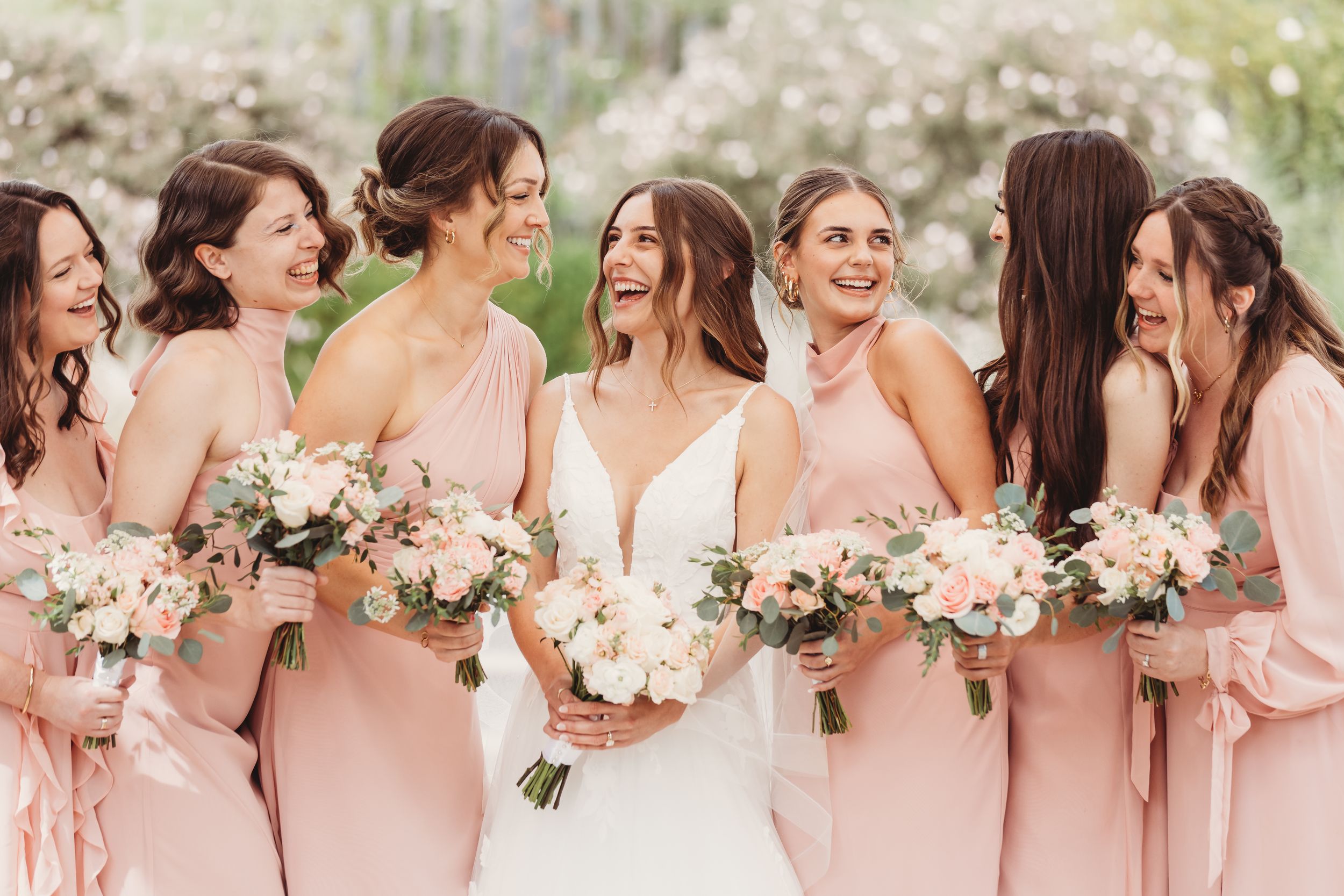 Bride in white gown laughing with six bridesmaids in blush pink dresses holding floral bouquets outdoors.