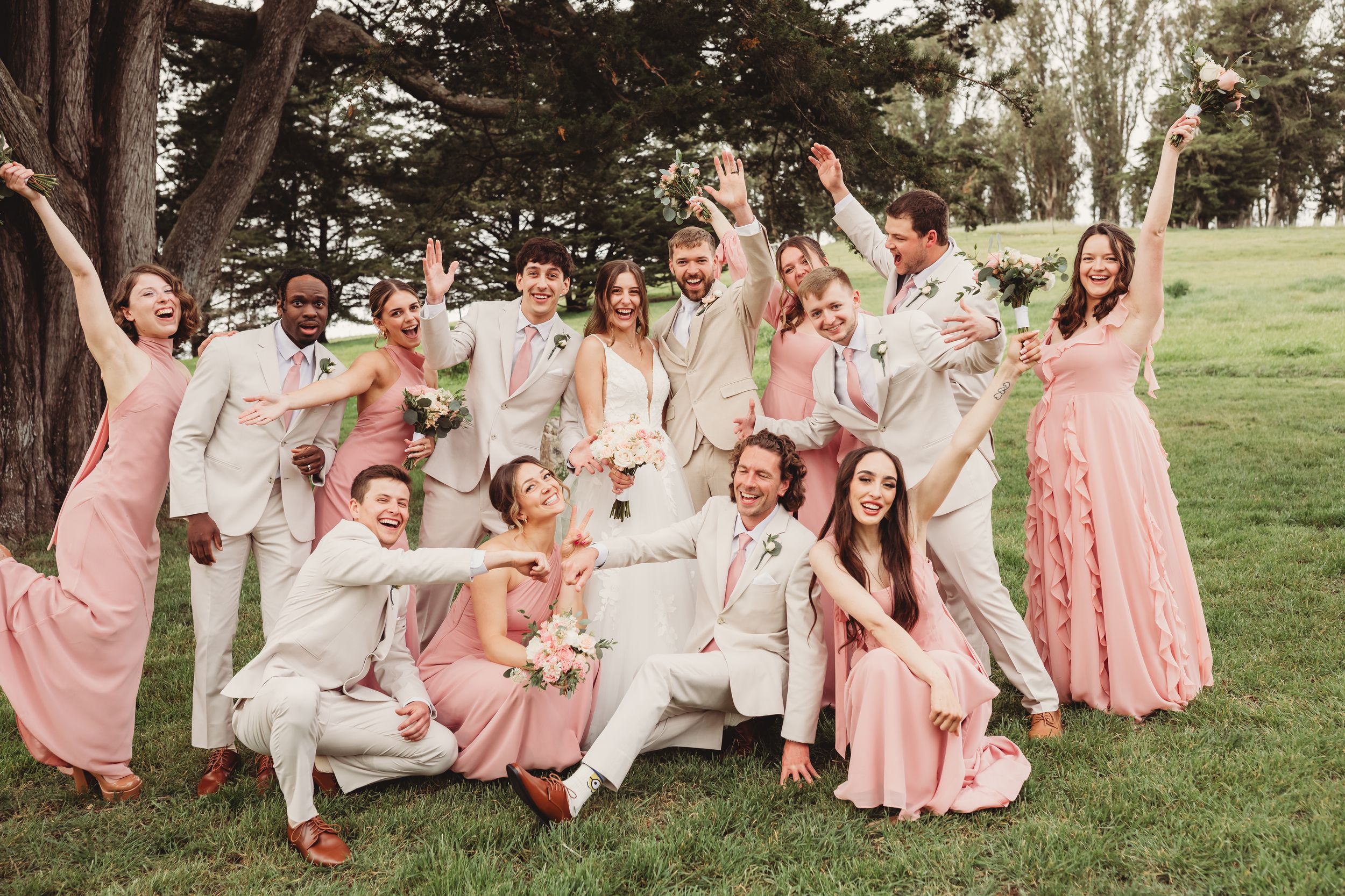 Wedding party in pink dresses and tan suits celebrating outdoors on green grass near trees.