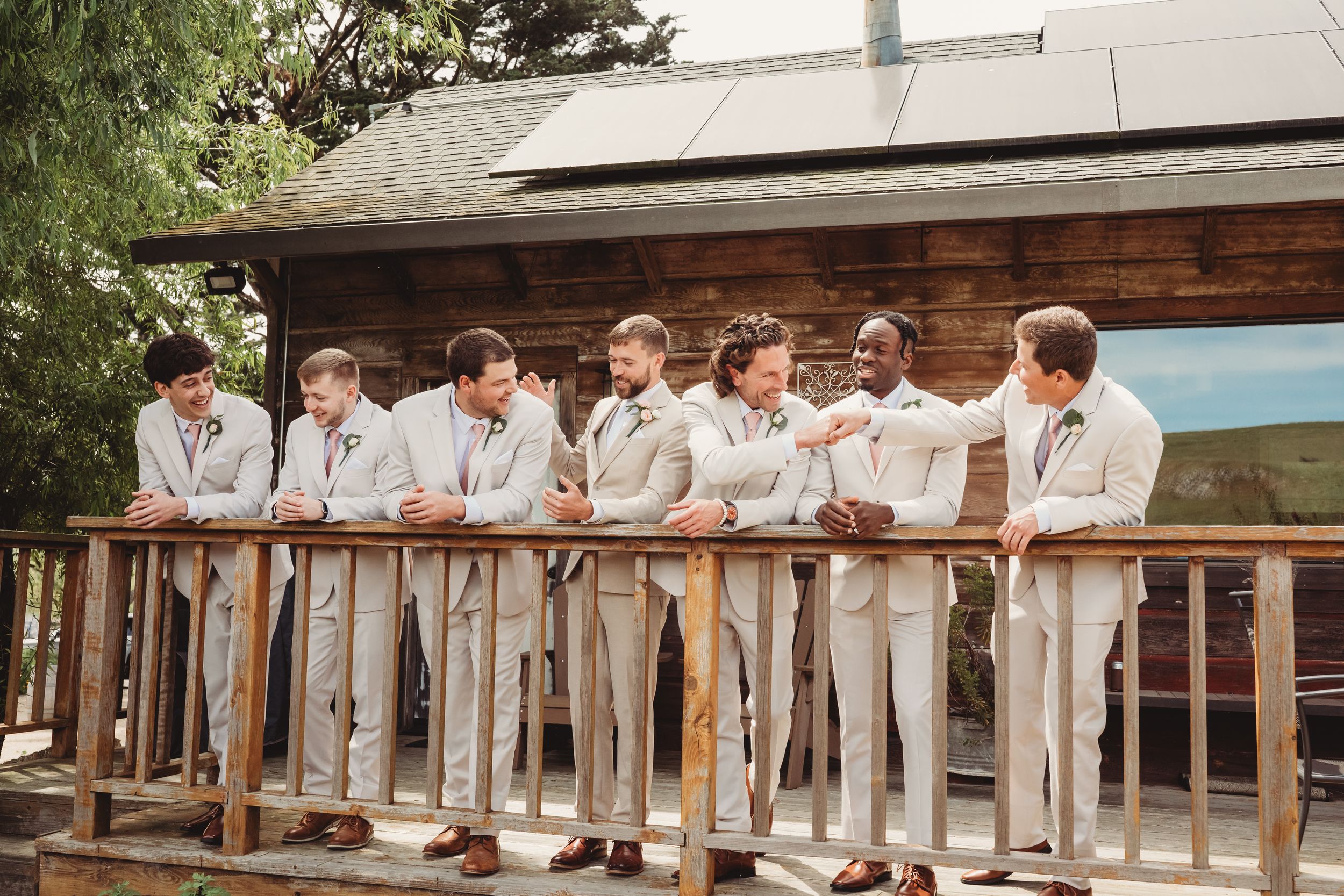Groomsmen in light gray suits posing together on a wooden deck outside a rustic venue on a wedding day.