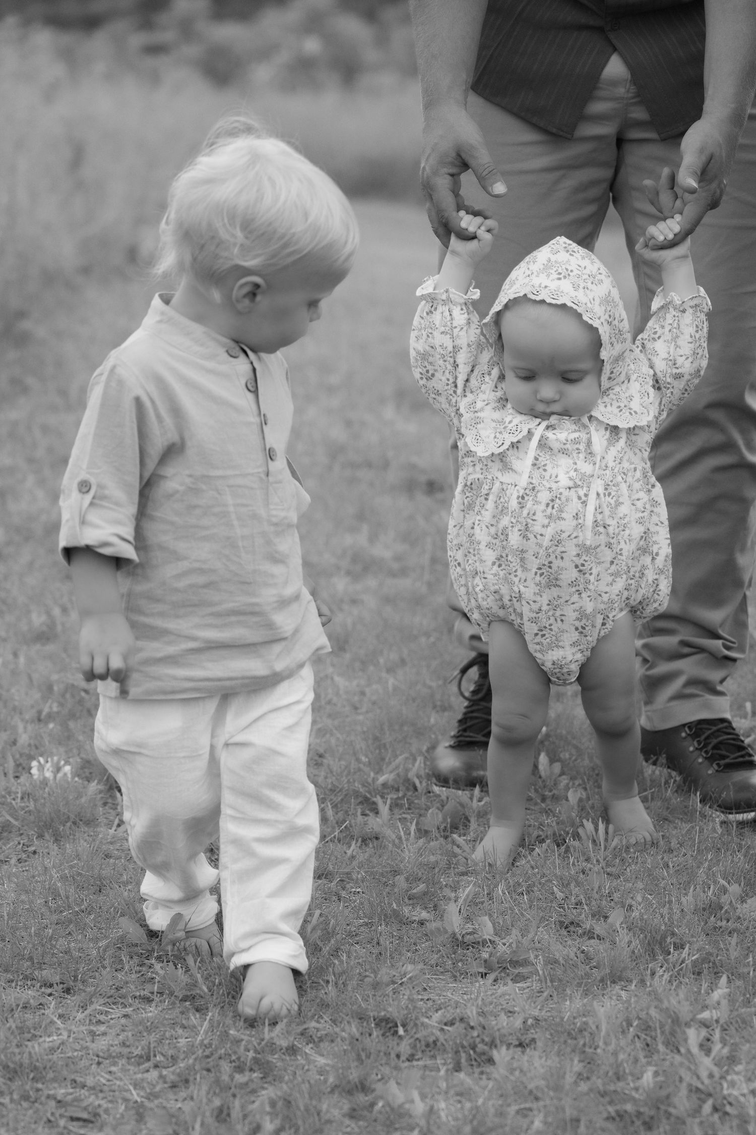 Candid of dad holding baby girl as she tries walking, her toddler brother watches during family photos in Hudson, WI.