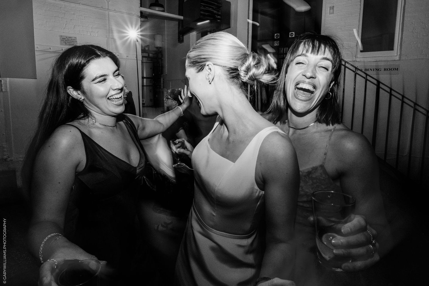 Three women laugh and dance together at a party in a black and white candid photo.