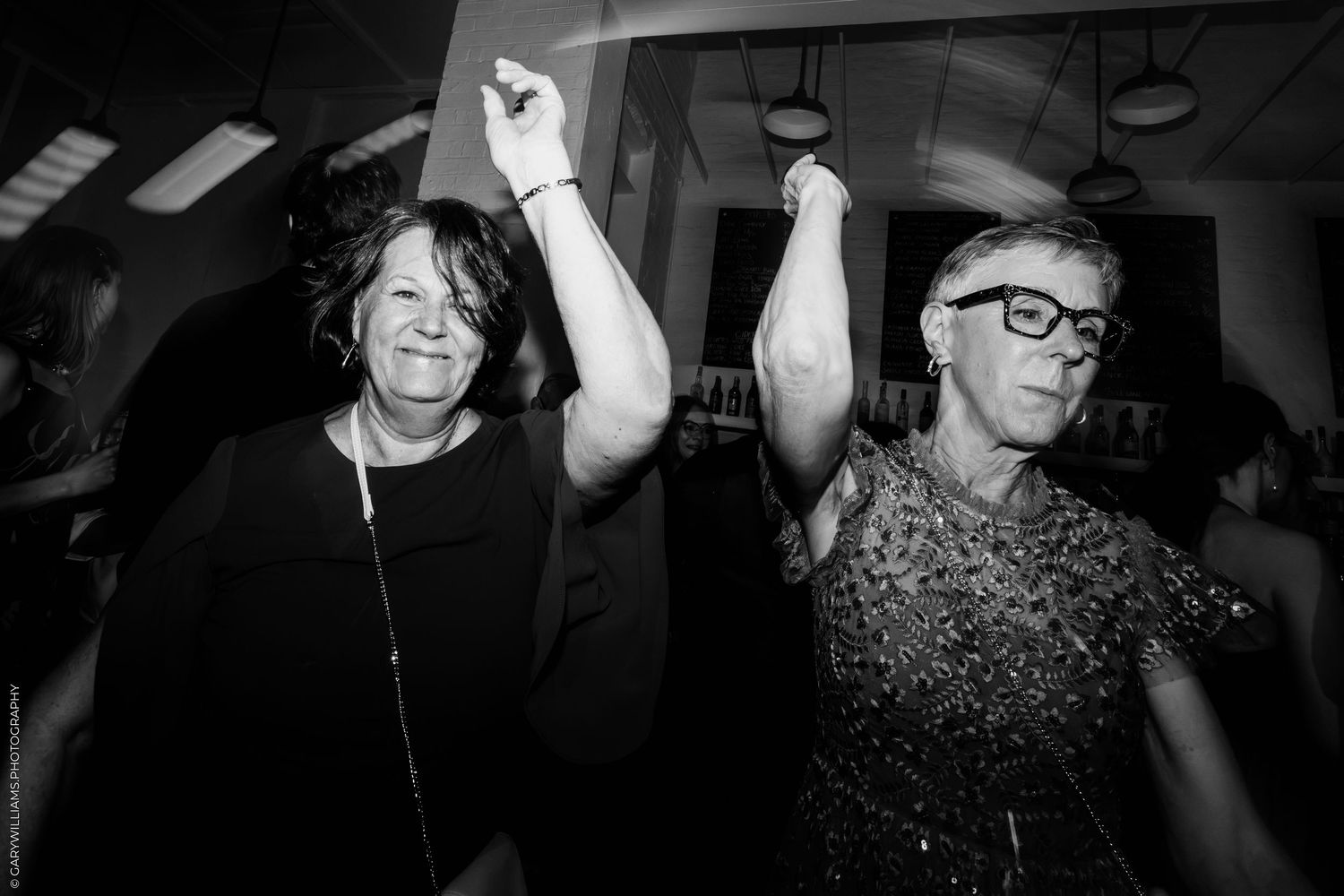 Two women dancing joyfully with arms raised at a party, captured in black and white photography.