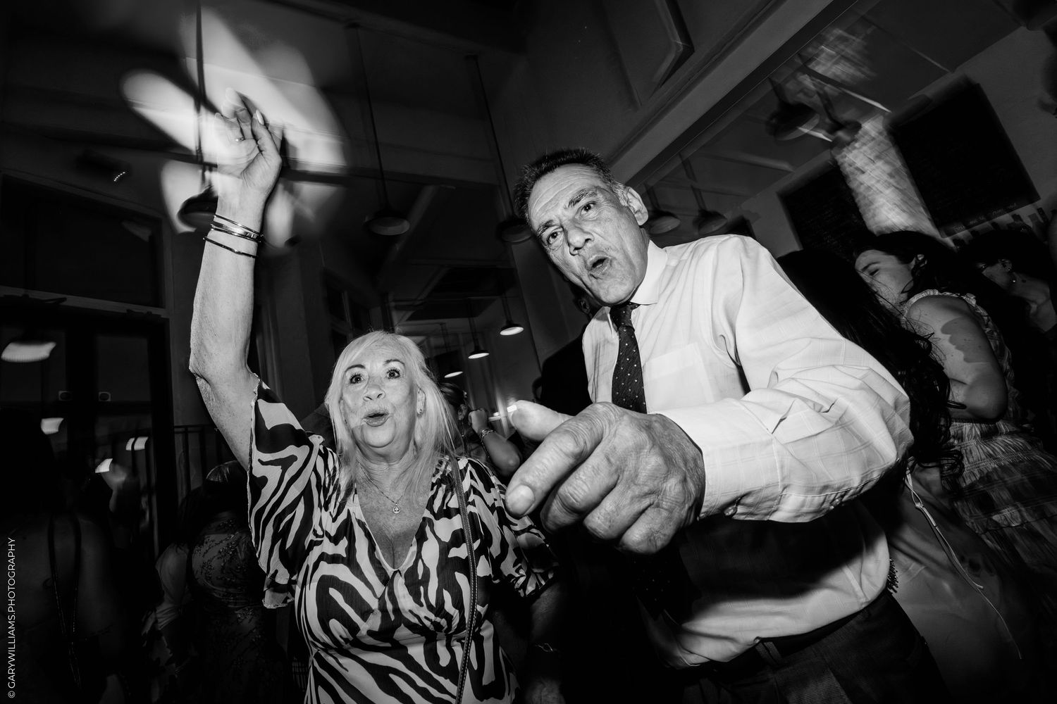 Black and white photo of two people dancing energetically at a party, woman raising arm, man pointing at camera.