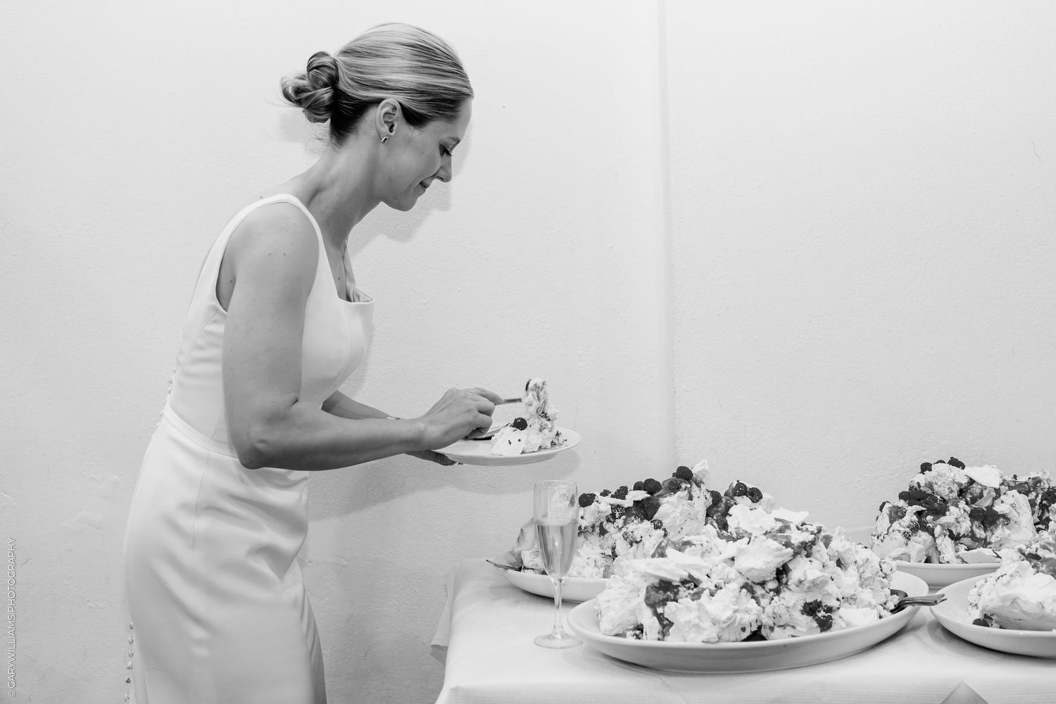 Bride in white dress cutting wedding cake at elegant reception table with floral desserts, black and white photo.