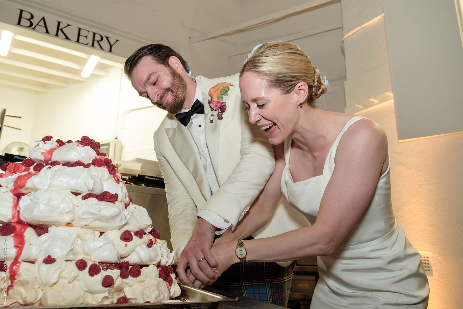 Bride and groom laughing while cutting white wedding cake with raspberries at bakery reception.