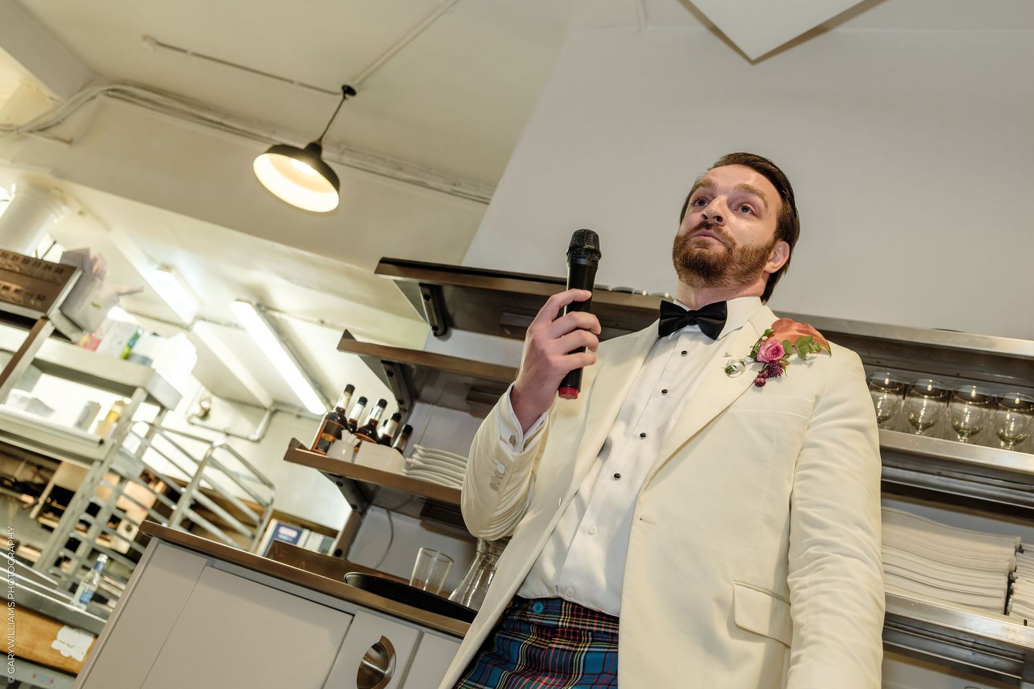 Groom in white jacket and tartan kilt giving speech with microphone.
