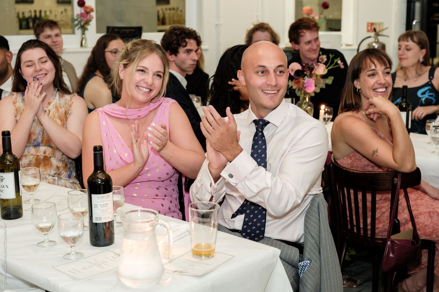 Guests clapping and smiling at a formal wedding reception dinner table with wine.