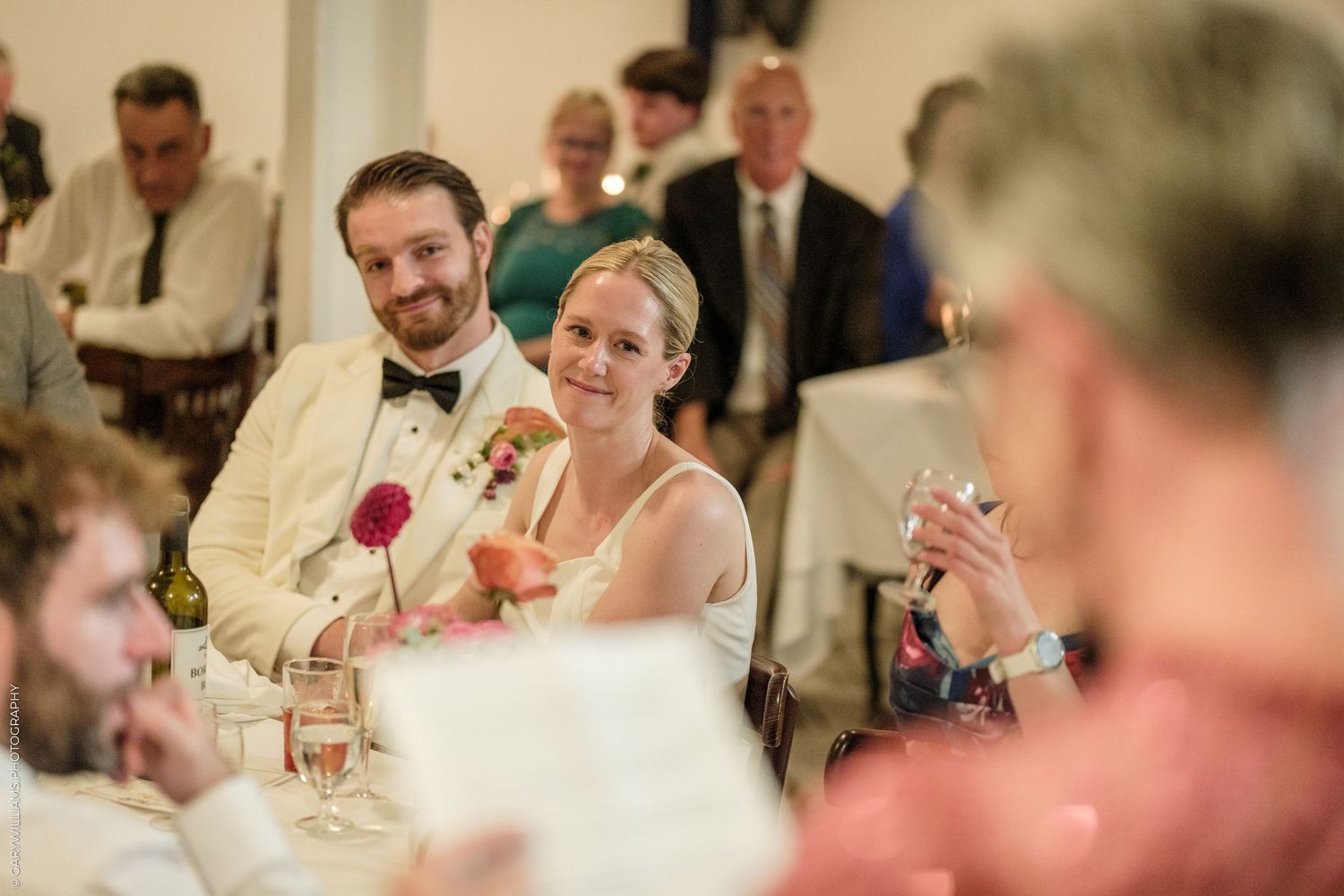 Bride and groom smiling during wedding reception toast, surrounded by guests in elegant venue.