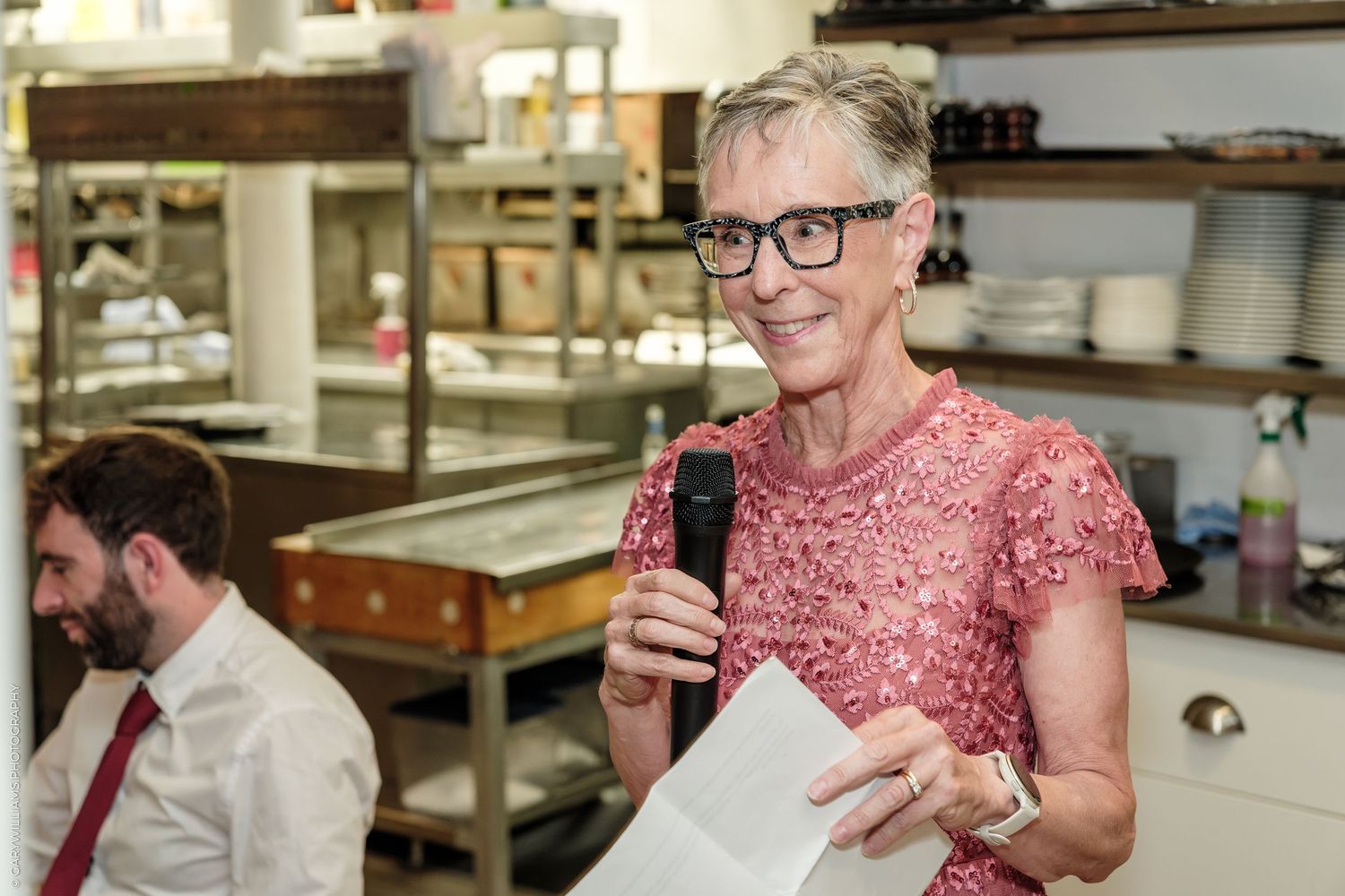 Elegant woman in pink lace dress speaks into microphone at upscale restaurant event.