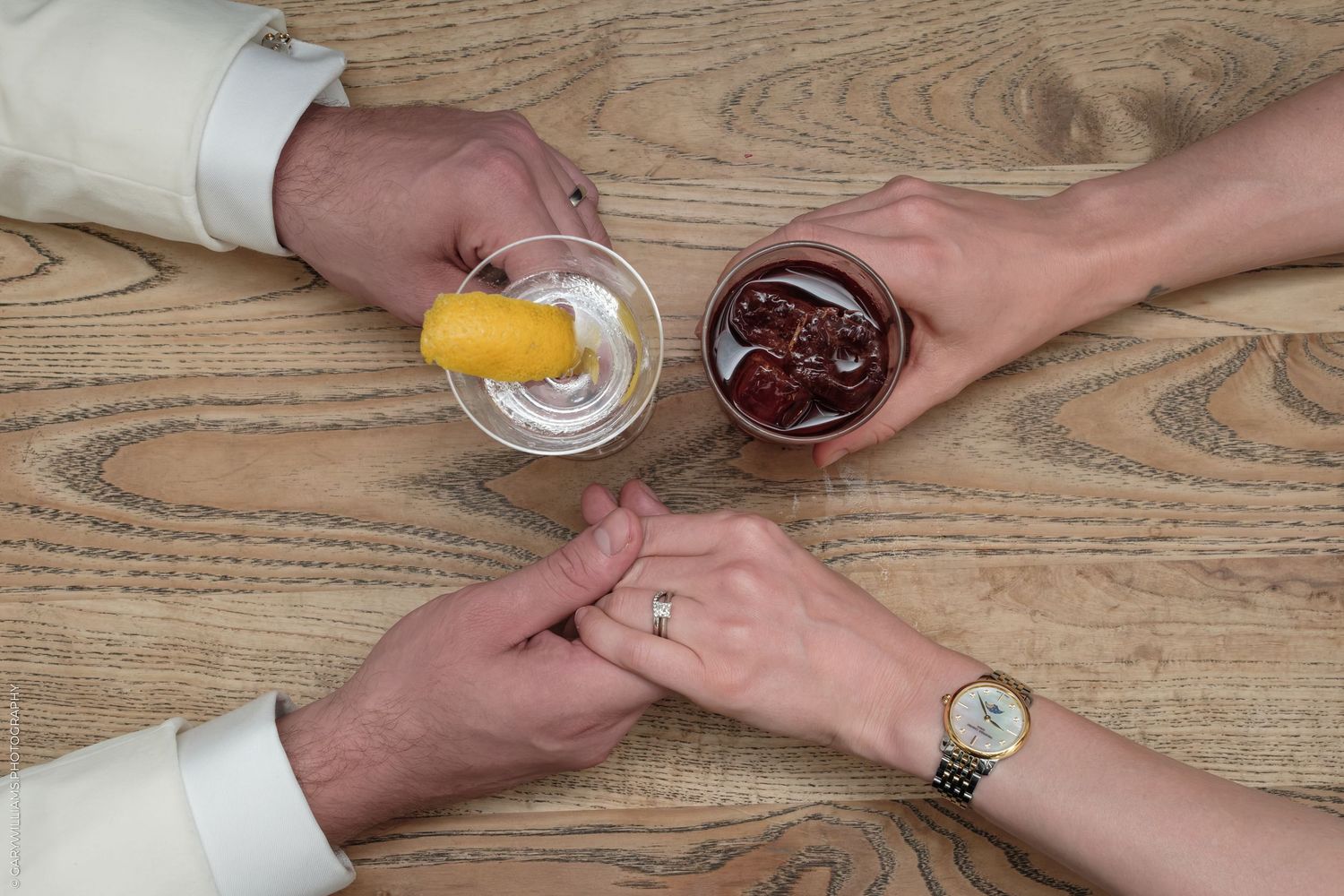 Couple holding hands on wooden table with cocktails, one drink garnished with lemon twist.