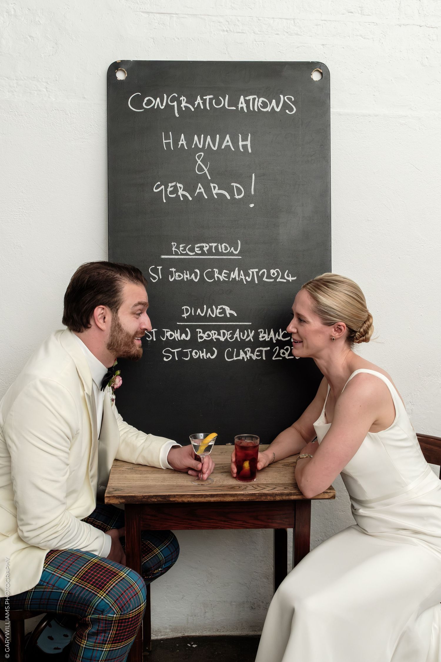 Newlyweds Hannah and Gerard sit at a small table in front of a congratulations chalkboard sign at their wedding reception.