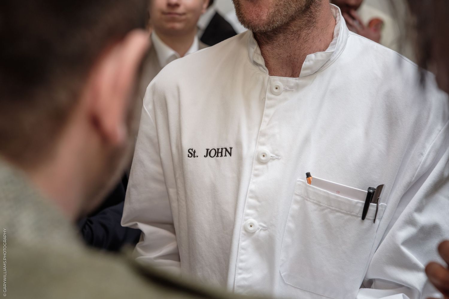 Chef in white St. John restaurant uniform with pen in pocket, standing among others in a professional kitchen setting.