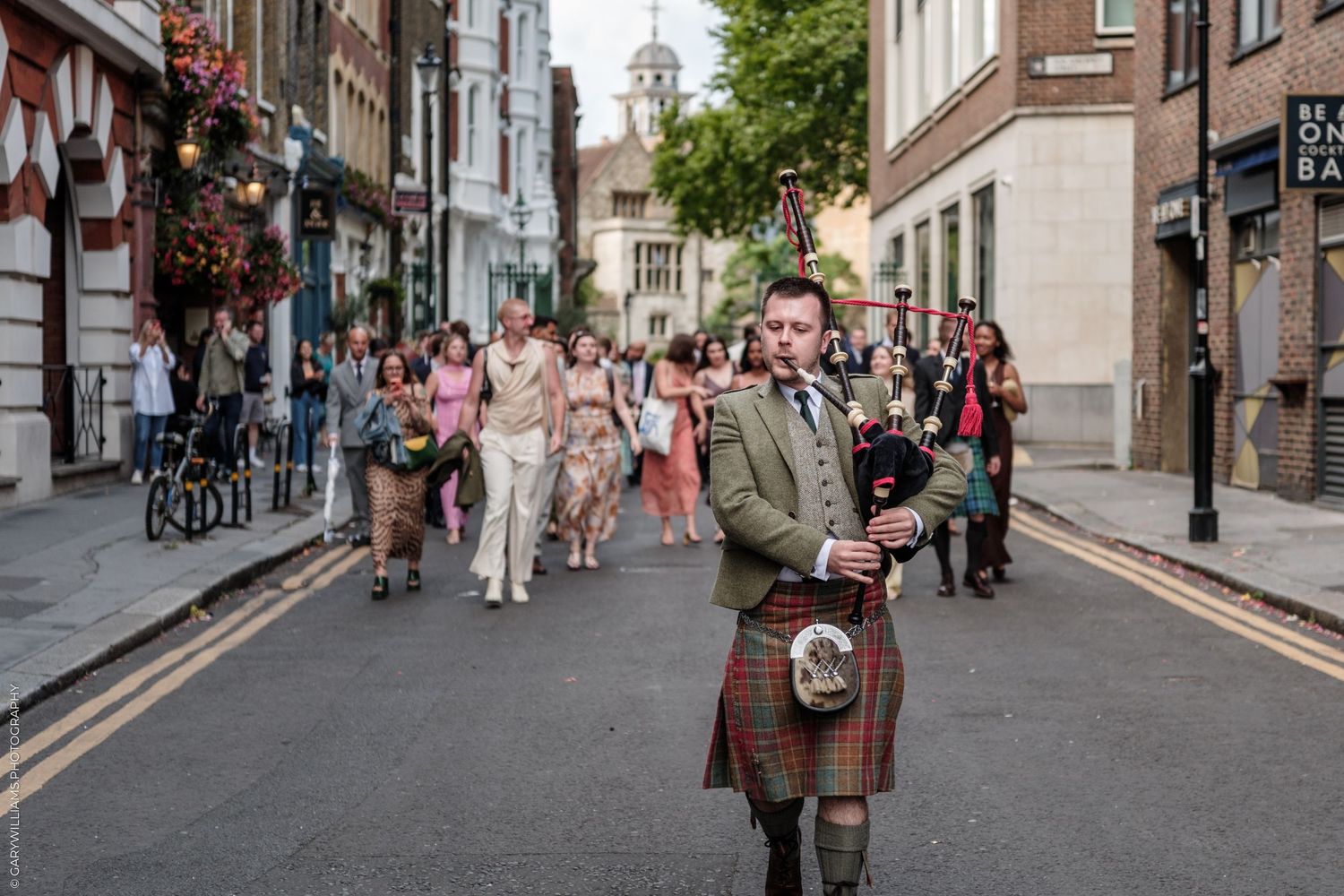 A bagpiper in tartan kilt leads a wedding procession down a cobblestone street in London.