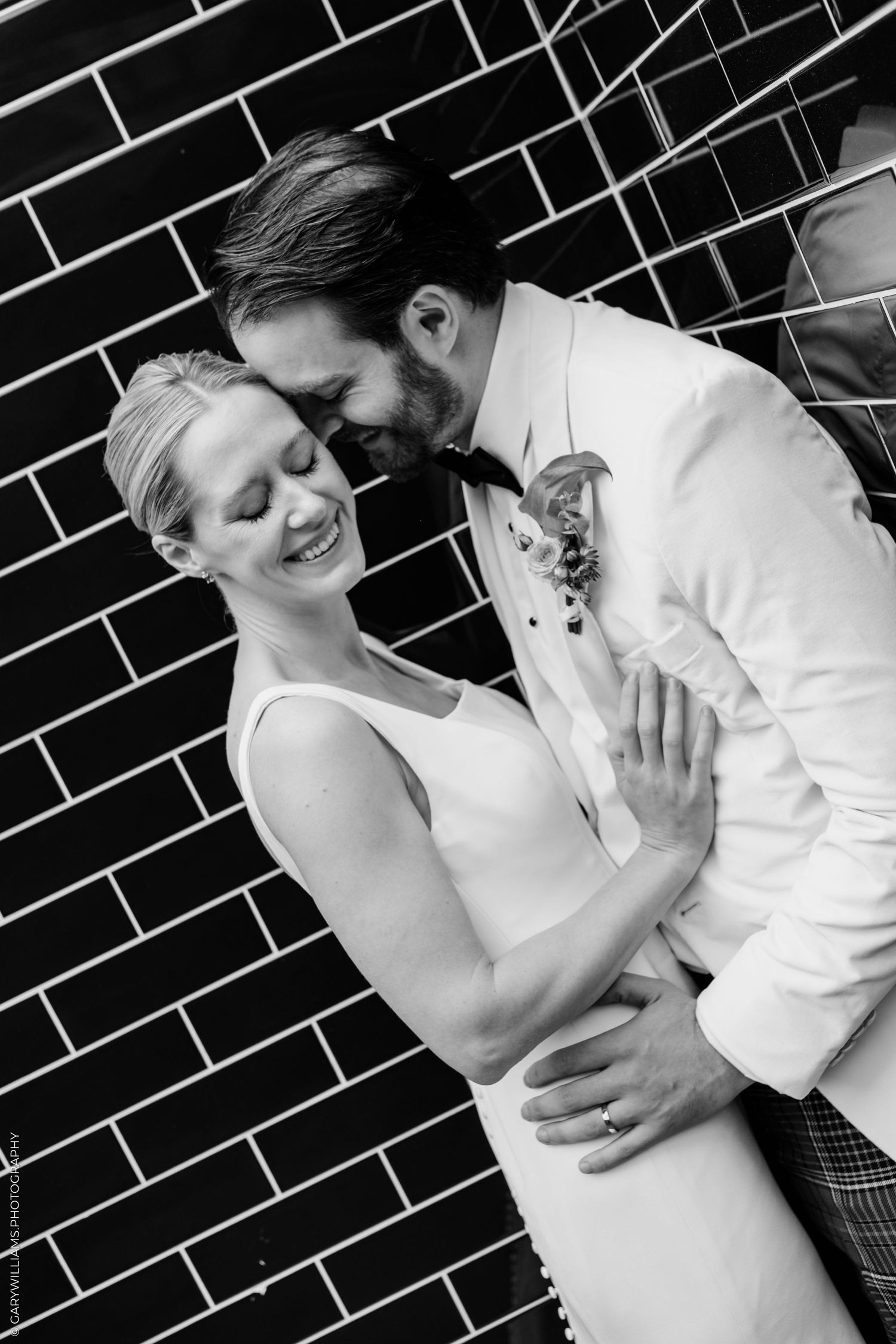 Black and white photo of a smiling bride and groom embracing against a brick wall on their wedding day.