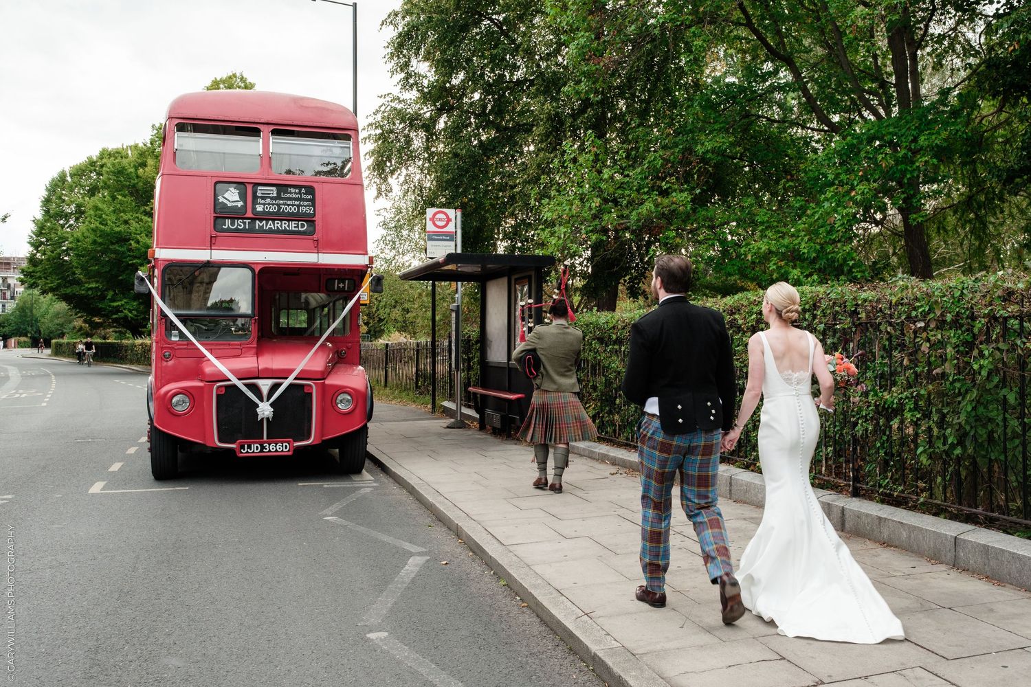 Bride in white gown and groom in Scottish kilt walking toward a red double-decker bus decorated with wedding ribbons.