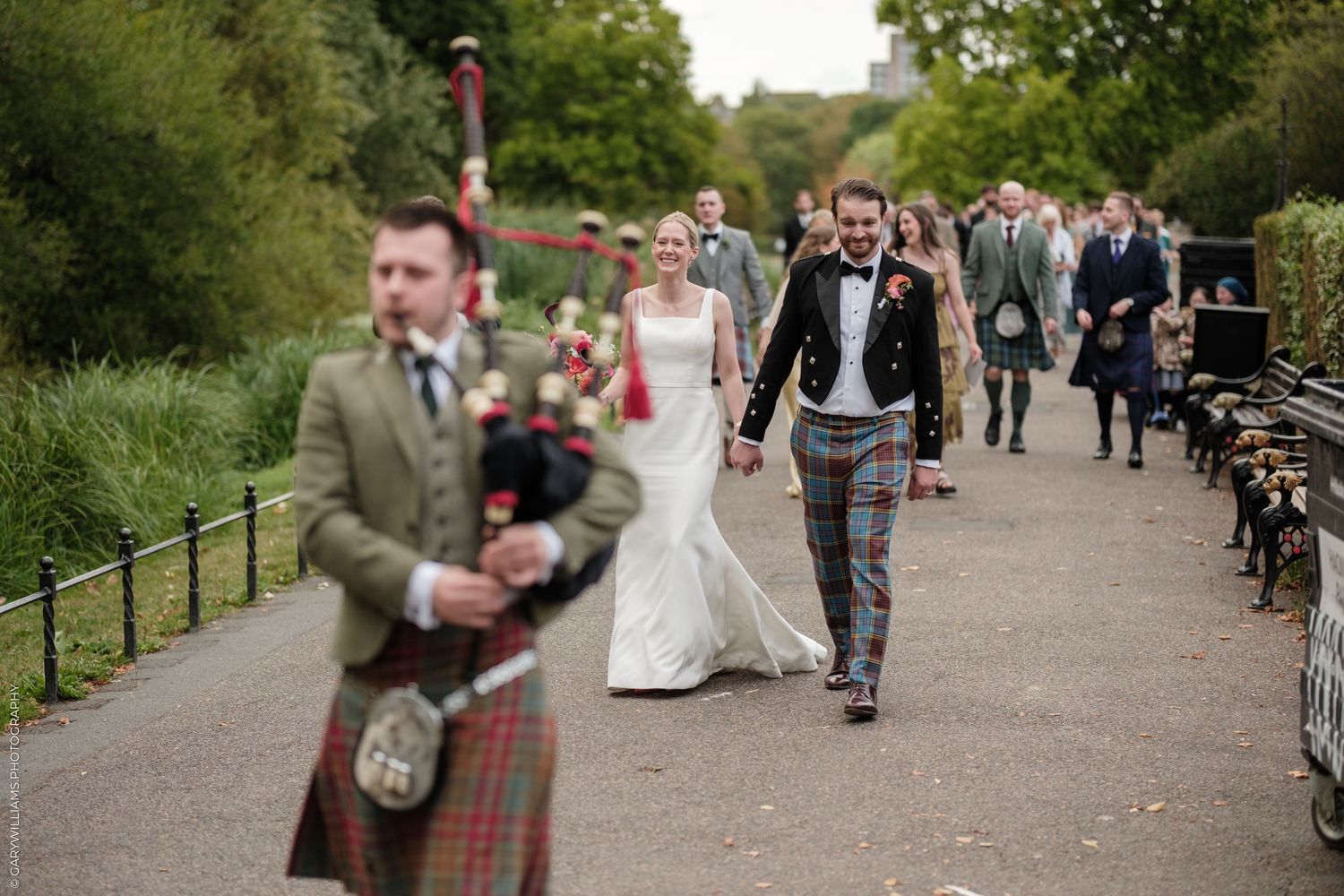 Scottish wedding procession with bagpiper leading bride and groom in tartan kilts along a tree-lined path.