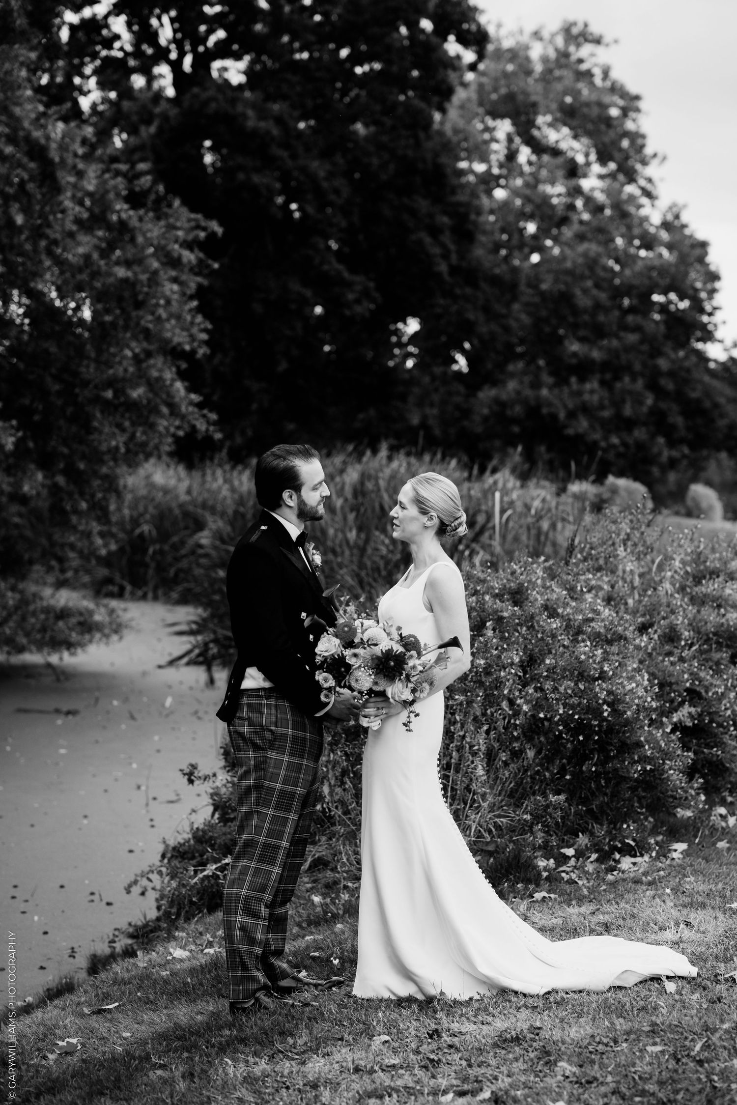 Black and white wedding photo of bride in gown and groom in tartan suit holding bouquet by a pond.