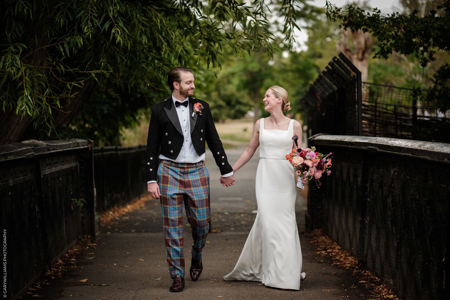 Bride and groom walking hand-in-hand on a bridge, groom in tartan kilt, bride in white gown holding pink bouquet.
