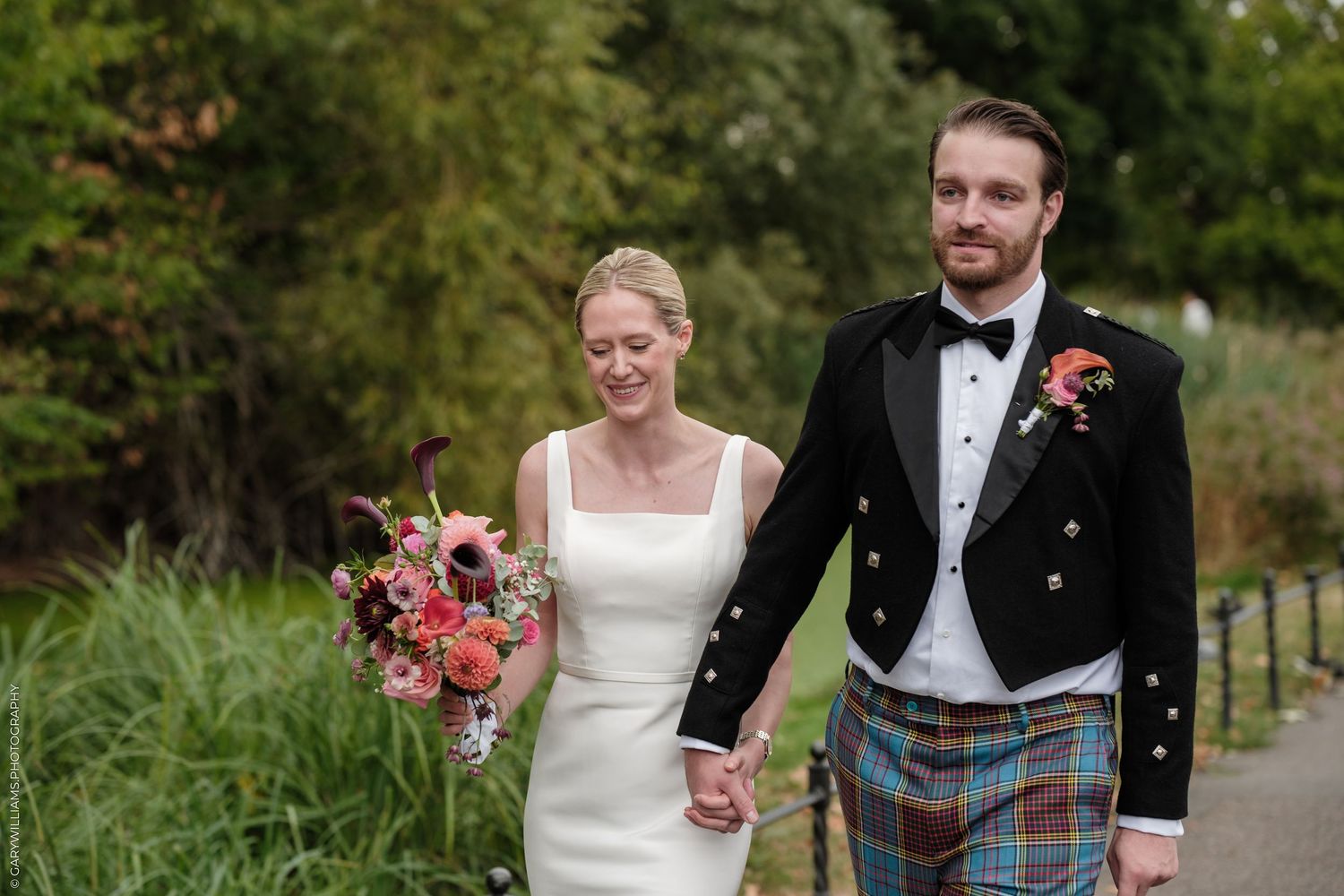 Bride and groom walk hand-in-hand, she holds a pink bouquet, he wears a tartan kilt and black jacket.