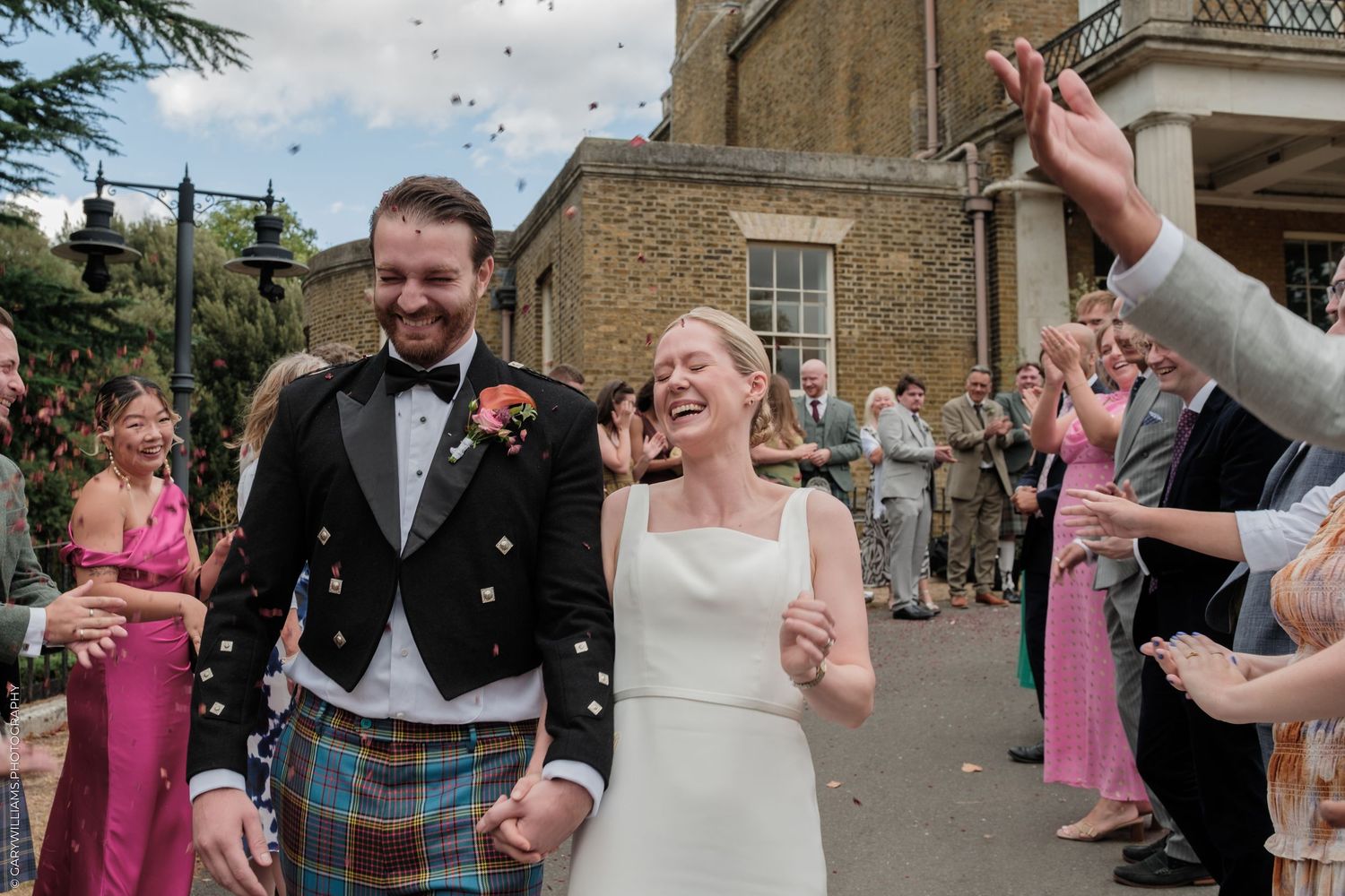 Joyful bride and groom walk through confetti shower outside elegant venue, groom wearing tartan kilt.