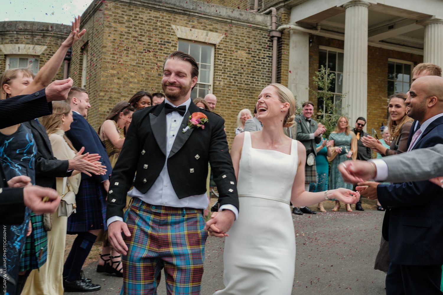 Joyful newlyweds walk through confetti shower, groom in tartan kilt, bride in white dress, wedding guests celebrating outdoors.