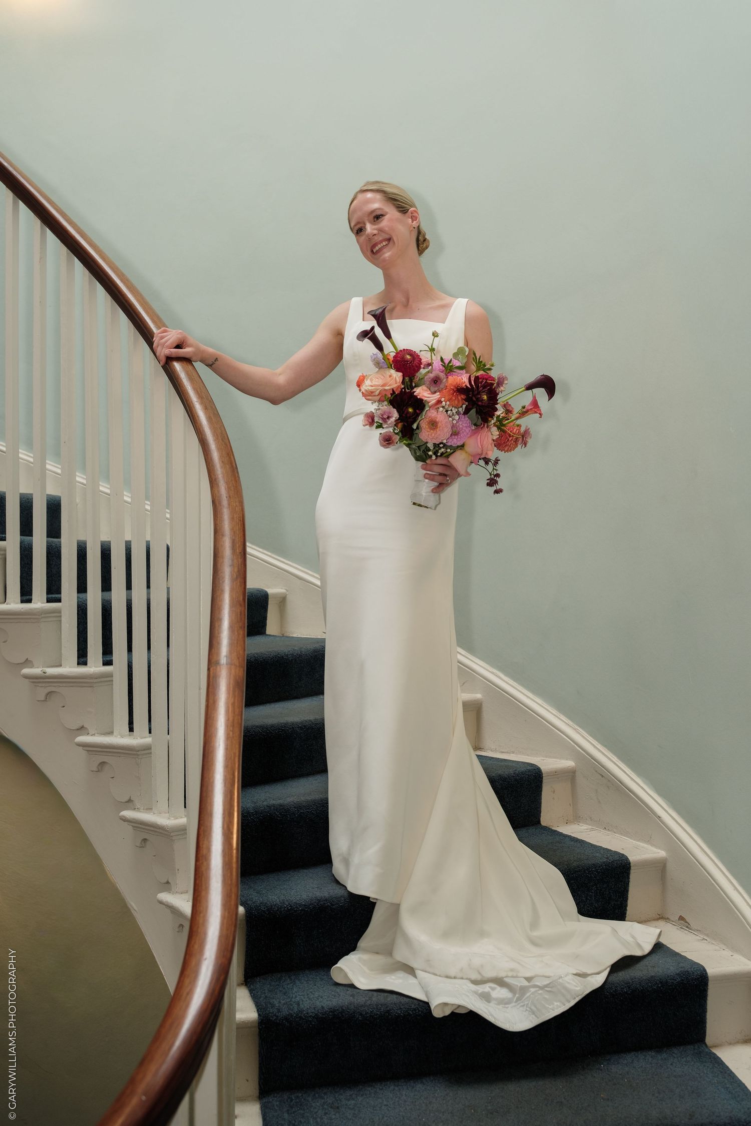 Bride in white gown holding colorful bouquet stands on curved staircase with dark carpet and wood railing.