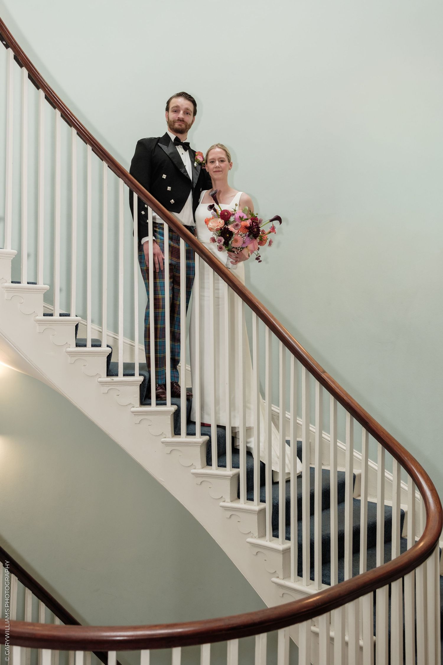 Bride and groom pose on elegant curved staircase, she holds colorful bouquet, he wears tartan kilt and black jacket.